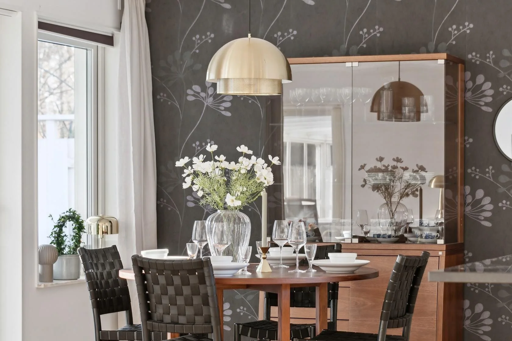 A dining room with a round wooden table set with white bowls, glasses, and a large vase with white flowers. There are four black woven chairs around the table and a window with white curtains on the left. A gold pendant light hangs above the table, a