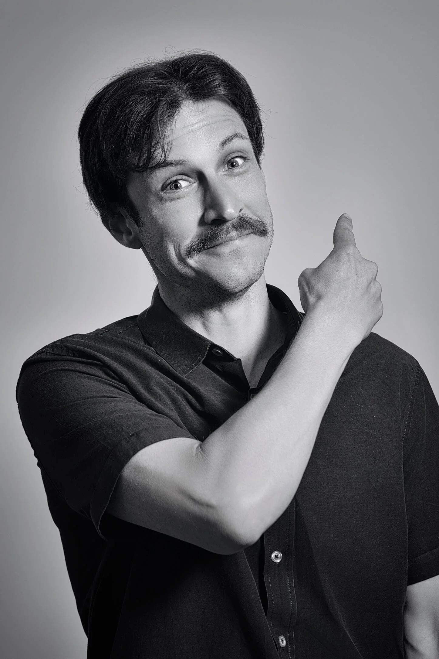 A black-and-white portrait of a man with dark hair and a mustache, smiling and pointing his right index finger upward, wearing a short-sleeved shirt, against a plain background.