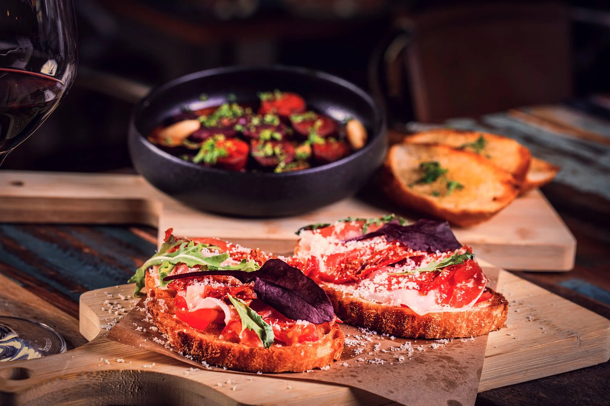 Open-faced sandwich with prosciutto, arugula, and Parmesan on toasted bread, garnished with purple basil leaves, served on a wooden board. In the background, a bowl of tomato salad with chopped herbs and two baked sweet potatoes, and a glass of red w