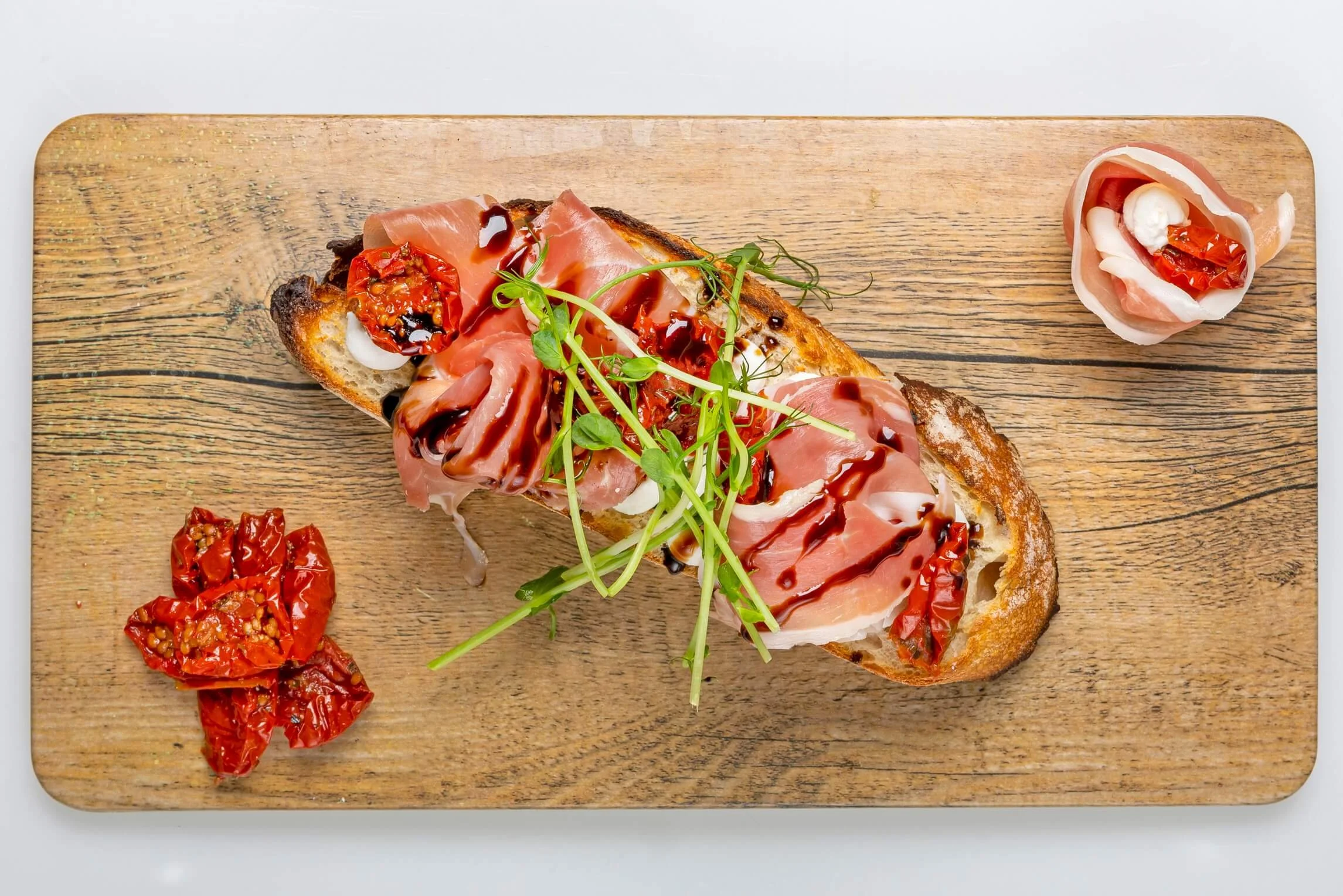 Open-faced bread topped with prosciutto, sun-dried tomatoes, microgreens, and balsamic glaze, served on a wooden board.
