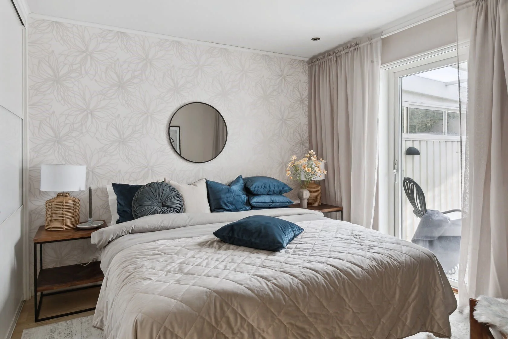 A cozy bedroom with a large bed covered in a beige quilt and blue pillows. A nightstand with a lamp and a vase with flowers is on the left. A round mirror hangs on an accent wall with subtle floral wallpaper. There are cream curtains and sliding glas