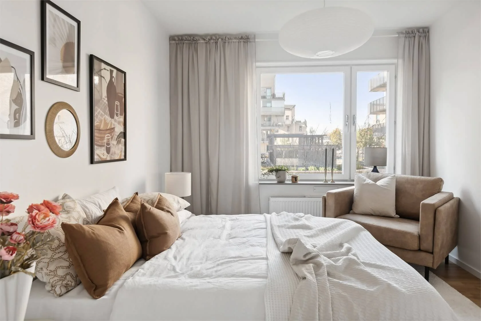 Bright bedroom with white bedding, multiple brown and beige pillows on the bed, beige armchair with pillow near window, framed art on white wall, curtains, window with city view, and decorative items on windowsill.