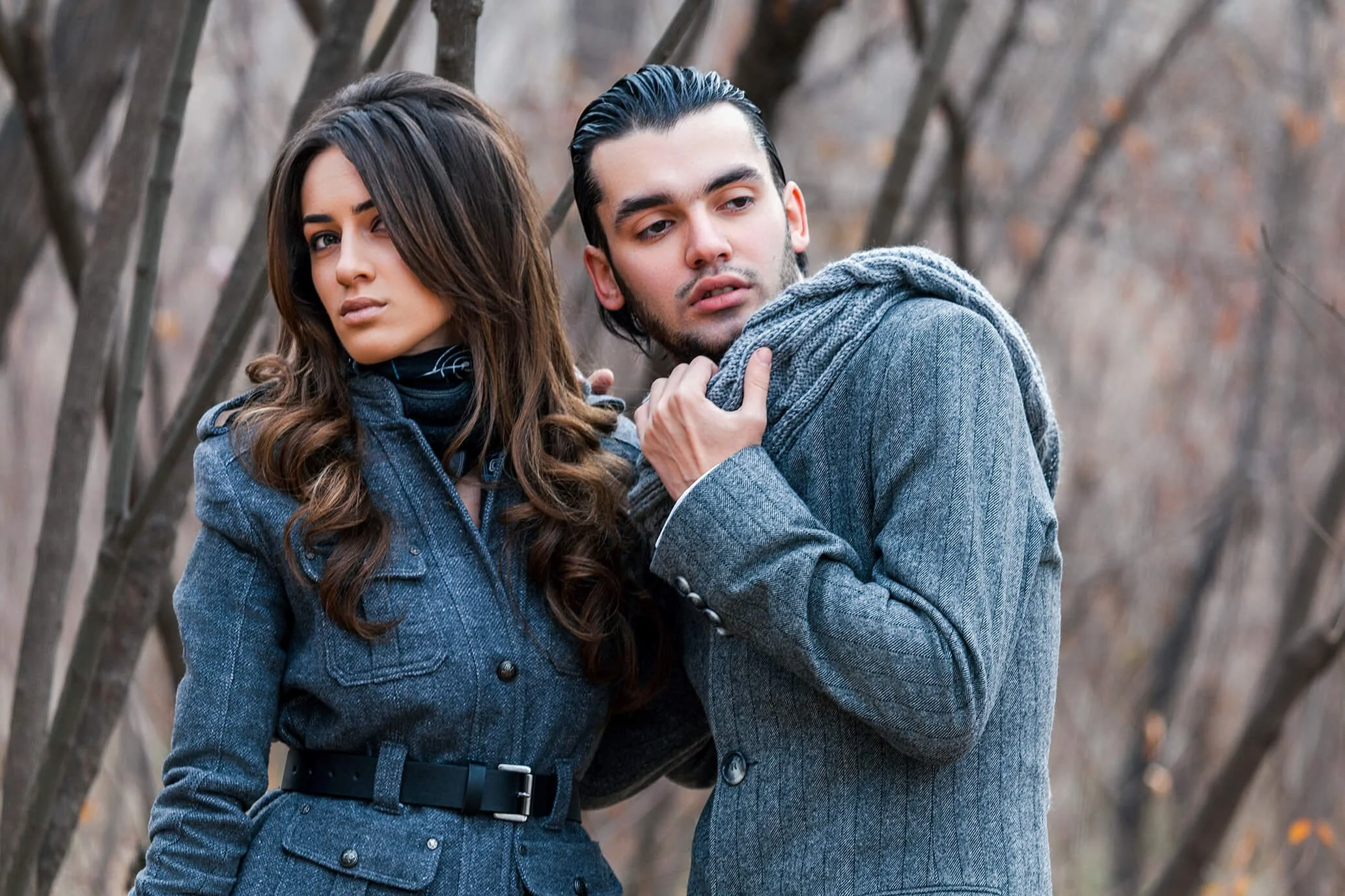 A man and a woman in stylish winter clothing standing outdoors among leafless trees, with the woman winking and the man looking away, both displaying serious expressions.