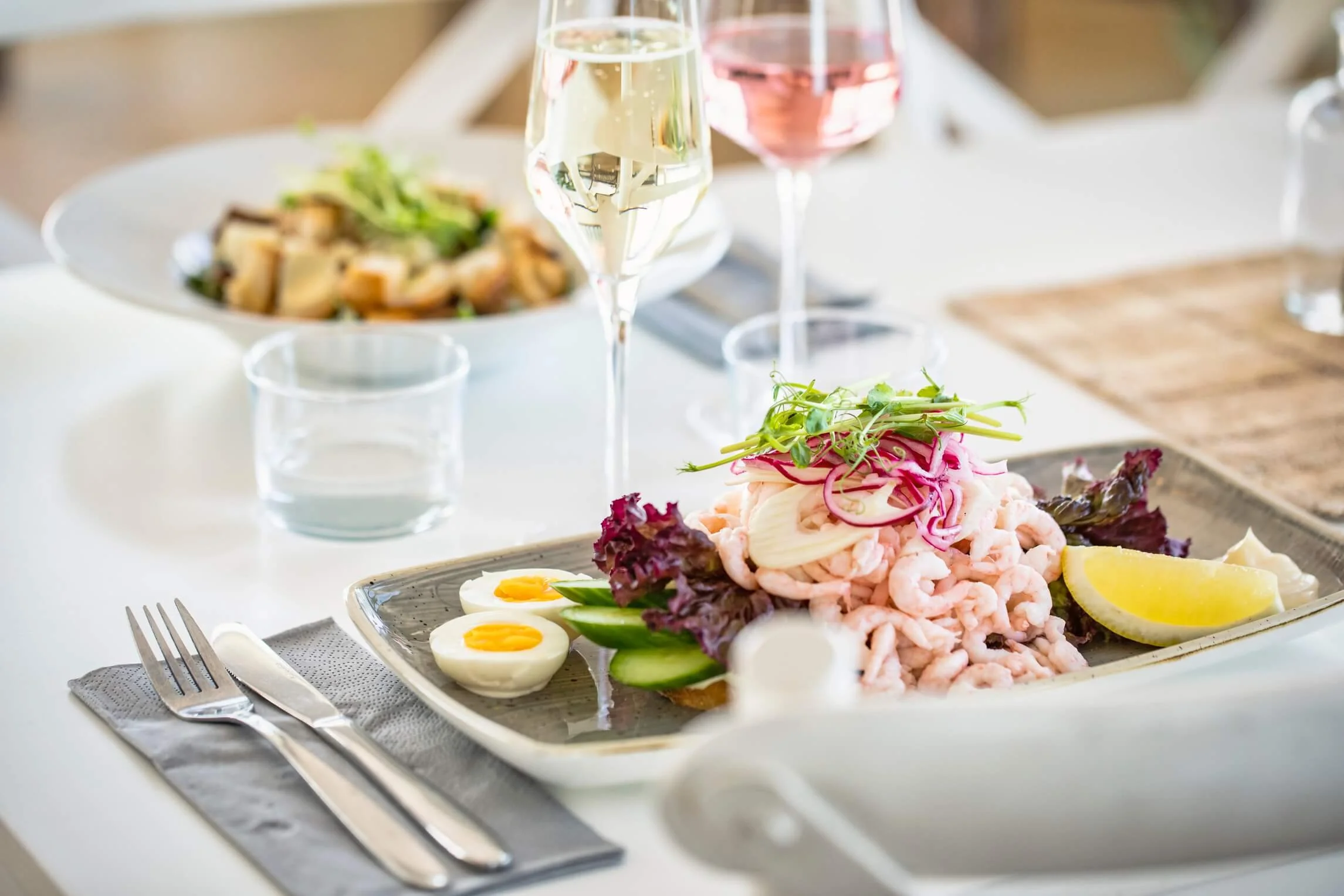 A plate of seafood salad with shrimp, boiled eggs, cucumber slices, microgreens, and lemon wedges. In the background, there is a glass of white wine, a glass of pink beverage, and a bowl of salad with greens and croutons on a white tablecloth.