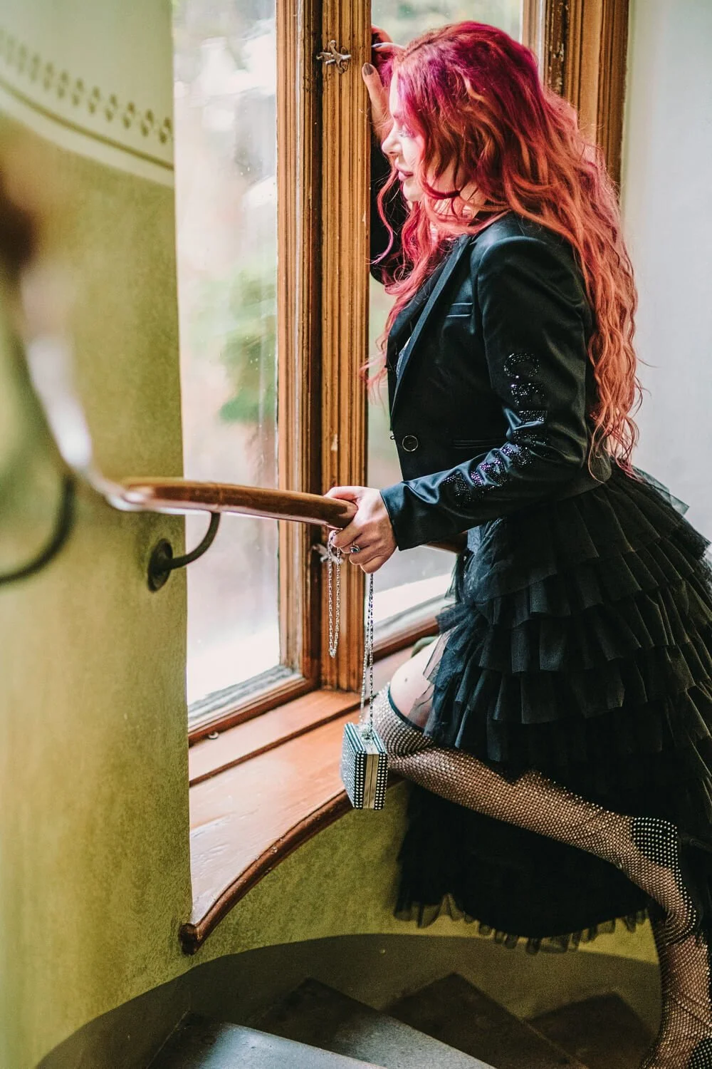 A woman with long red hair in a black jacket and layered skirt kneeling by a window, holding onto the window frame, with a chain-link handbag hanging from her wrist, indoors with green walls.