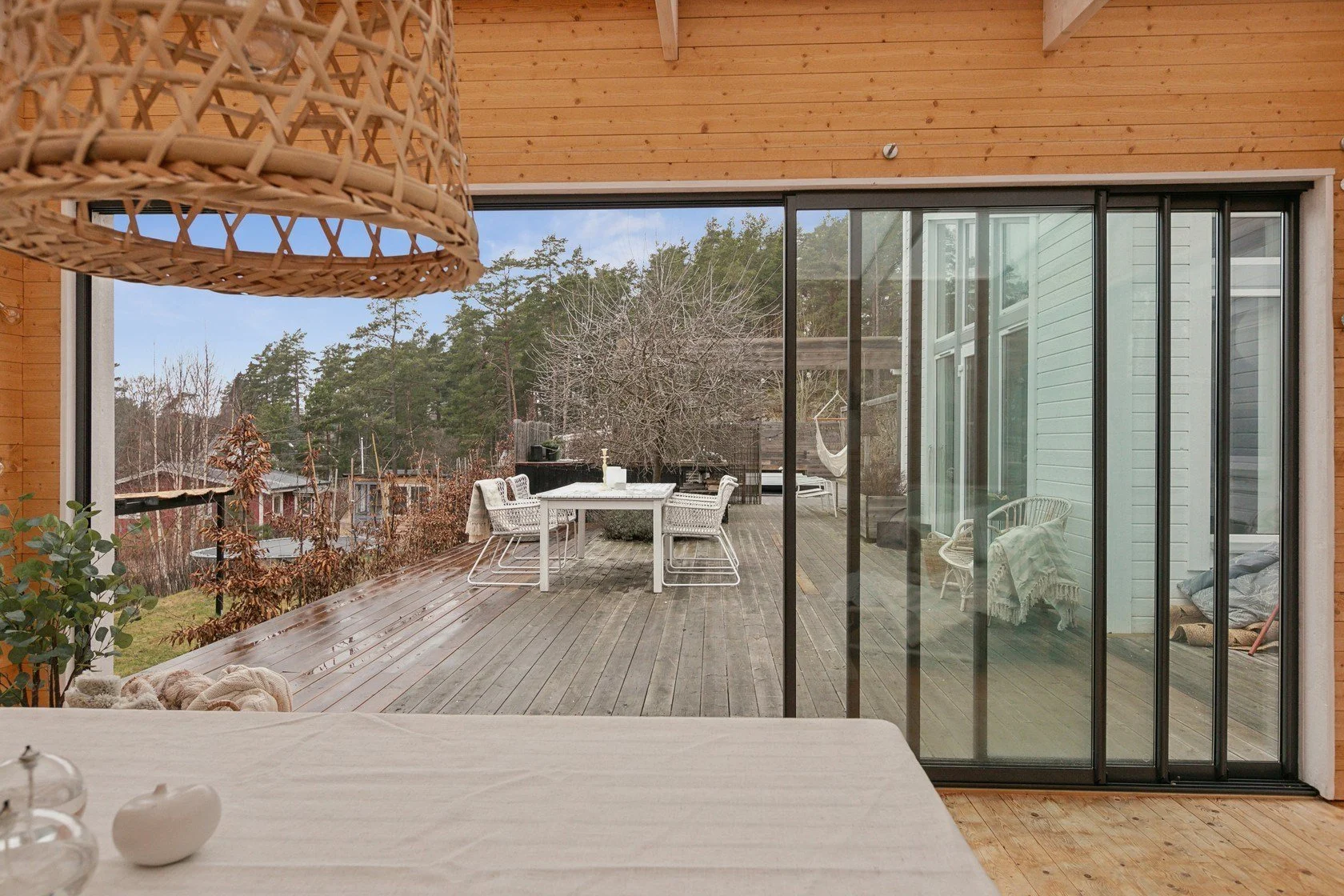 View of a wooden deck seen through large glass sliding doors from inside a house, with a white table and chairs on the deck, trees, and neighboring houses in the background.