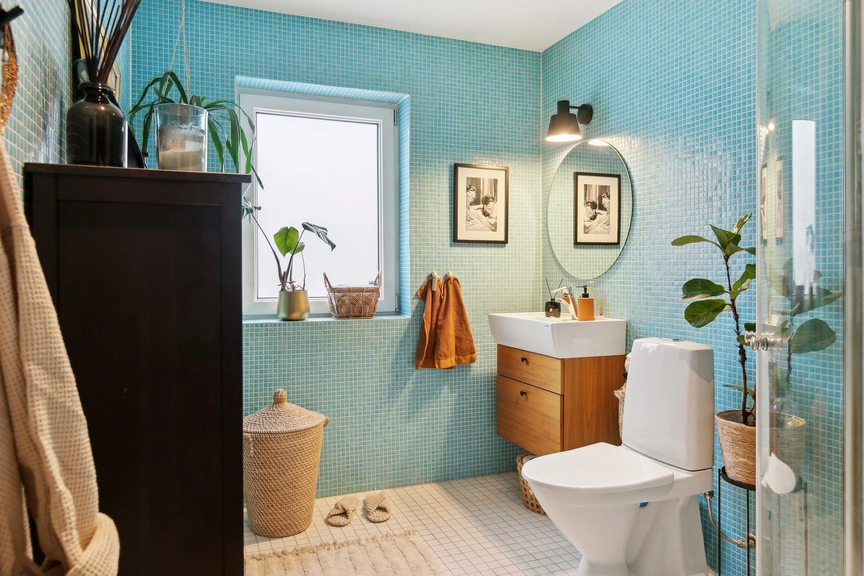 Bathroom with blue mosaic tile walls, a window with a potted plant, framed black and white photographs, a white sink with a wooden cabinet, a toilet, and various decorative plants and accessories.