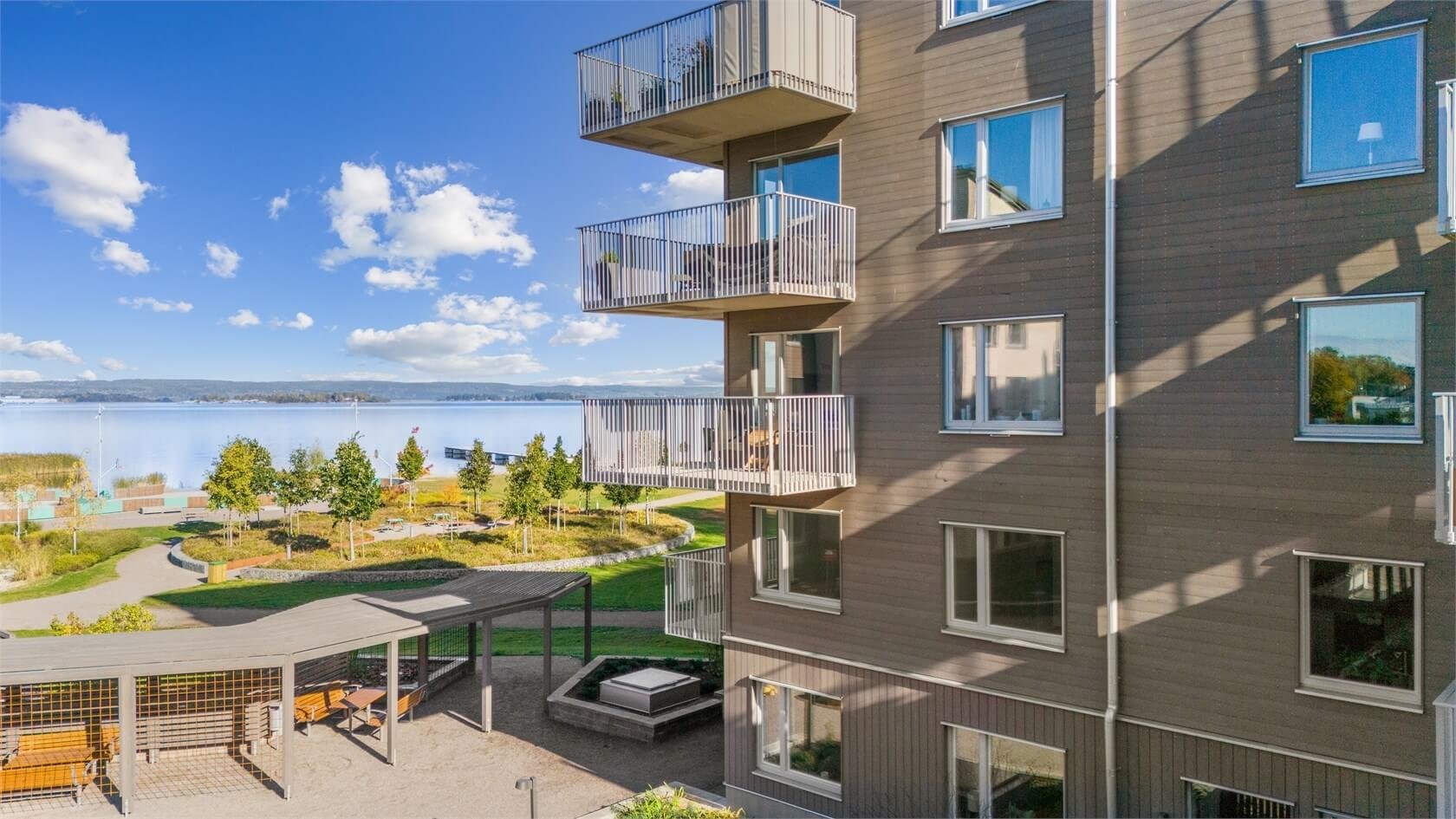 View of a multi-story residential building with balconies overlooking a waterfront area with paved walkways, greenery, and trees, under a partly cloudy sky.