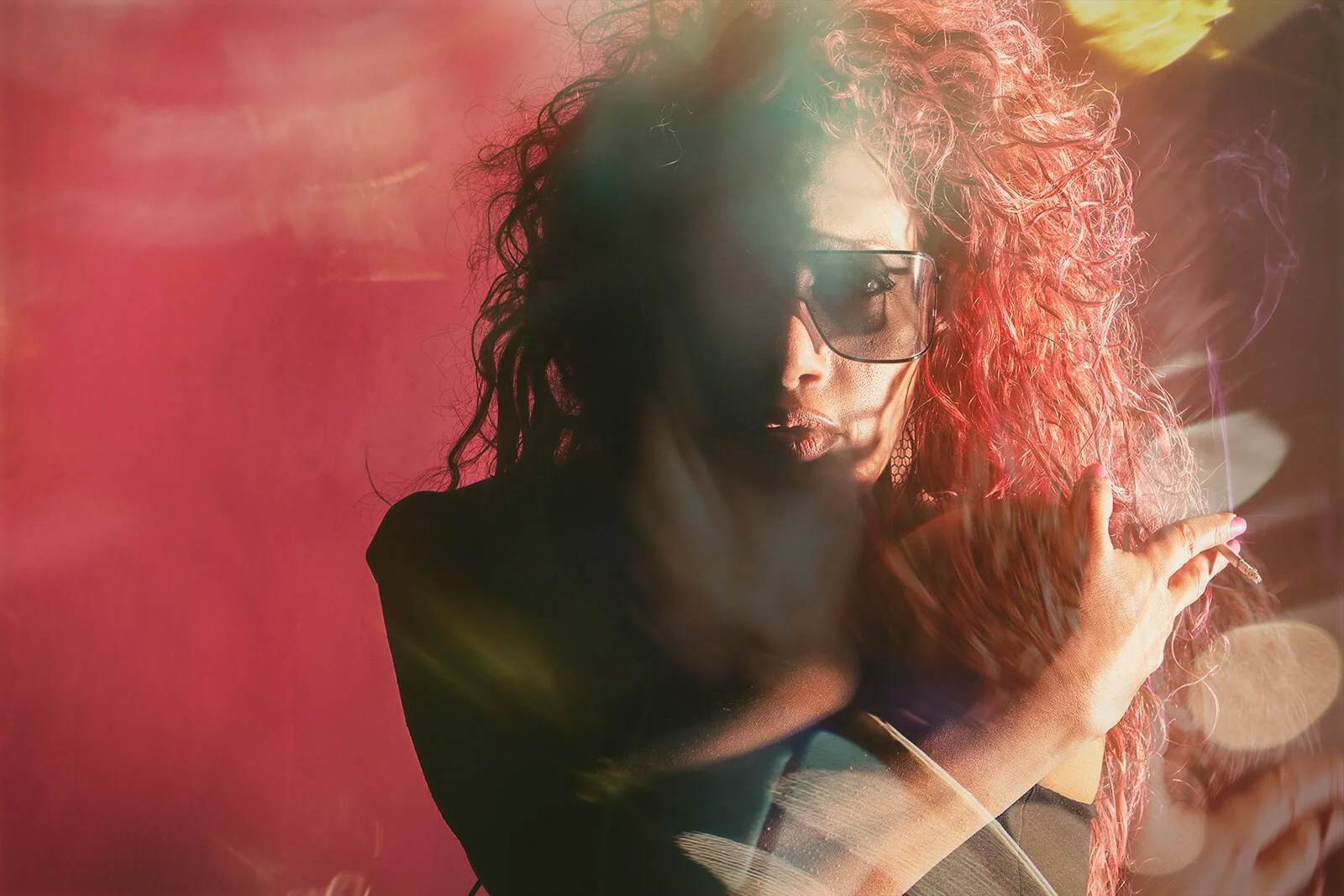 A woman with curly red hair wearing sunglasses smokes a cigarette against a pink background with light flare effects.