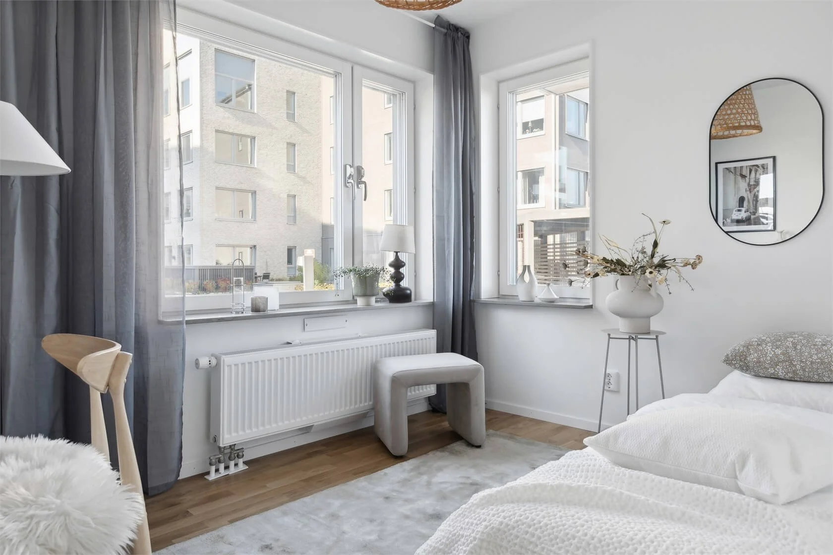 Bright bedroom with white walls, large windows, and minimalist decor. Contains a bed with white bedding, a white vase with dried flowers on a stand, a tufted ottoman, and decorative vases. A mirror and a lamp are also visible.