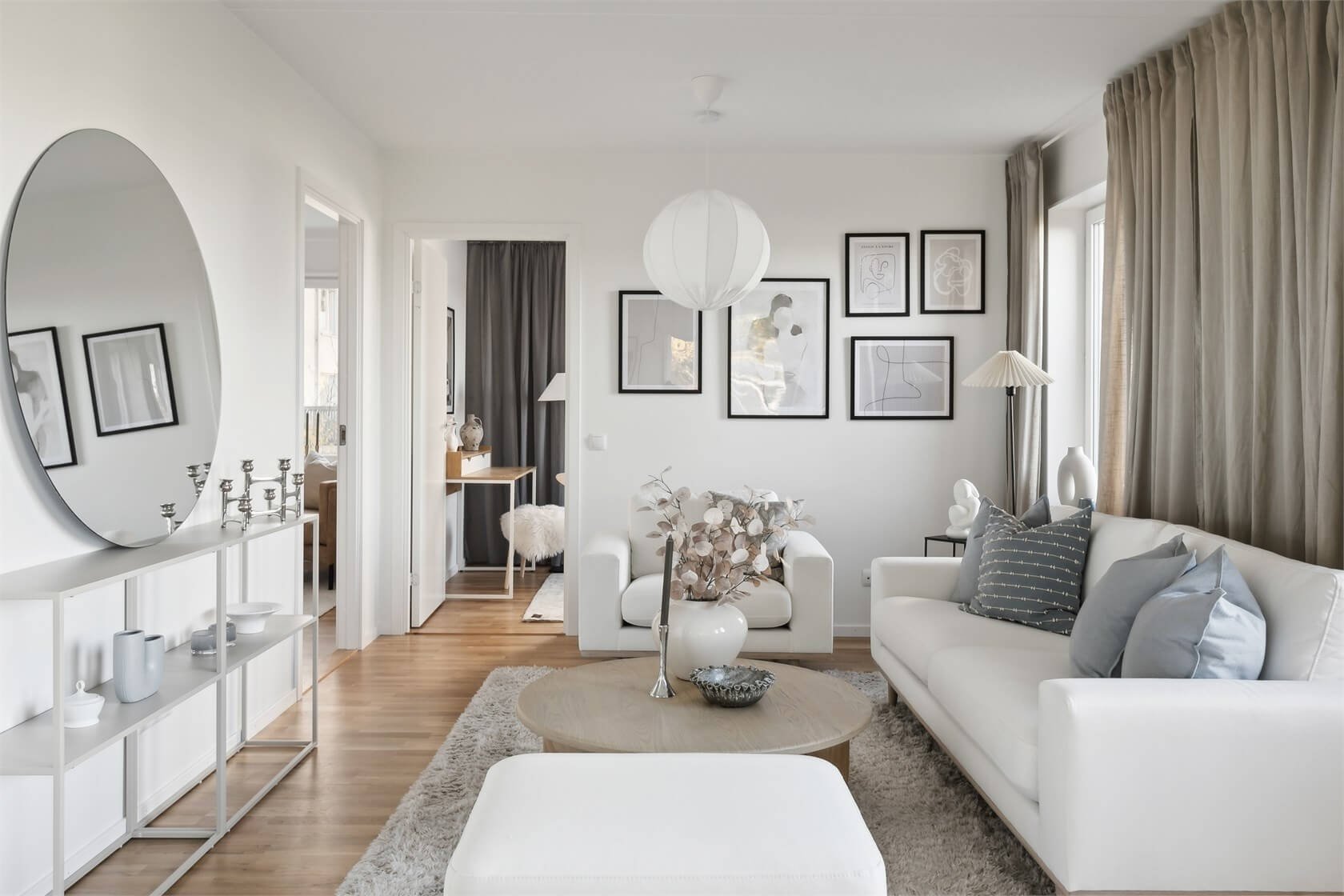 Minimalist living room with white sofa, armchair, wooden coffee table, black framed artwork, tall window with beige curtains, decorative vases, and a large round wall mirror.