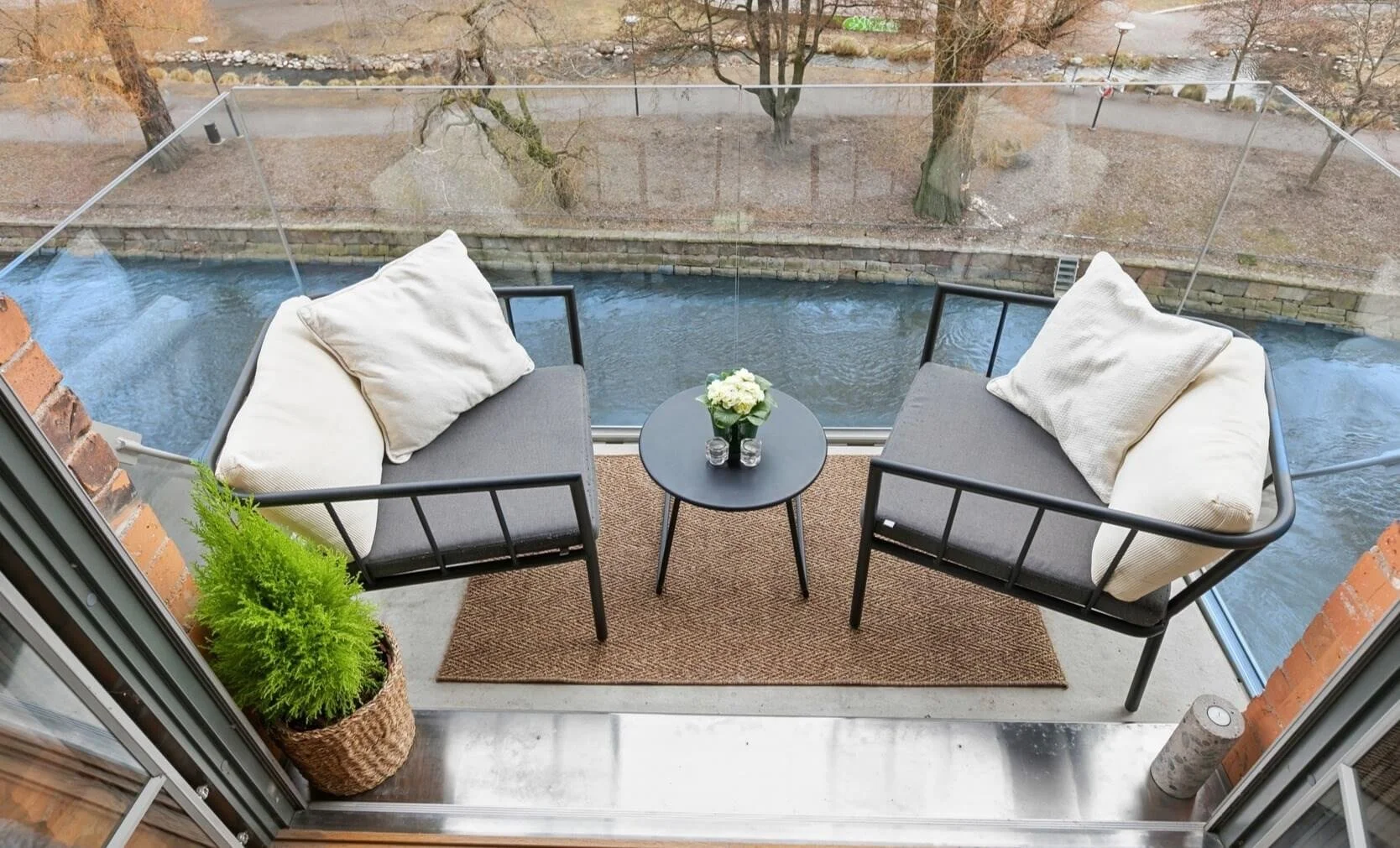 Balcony with two black metal chairs with gray cushions and white pillows, a small black table with a flower vase, outdoors view of river, trees, and pedestrian walkway