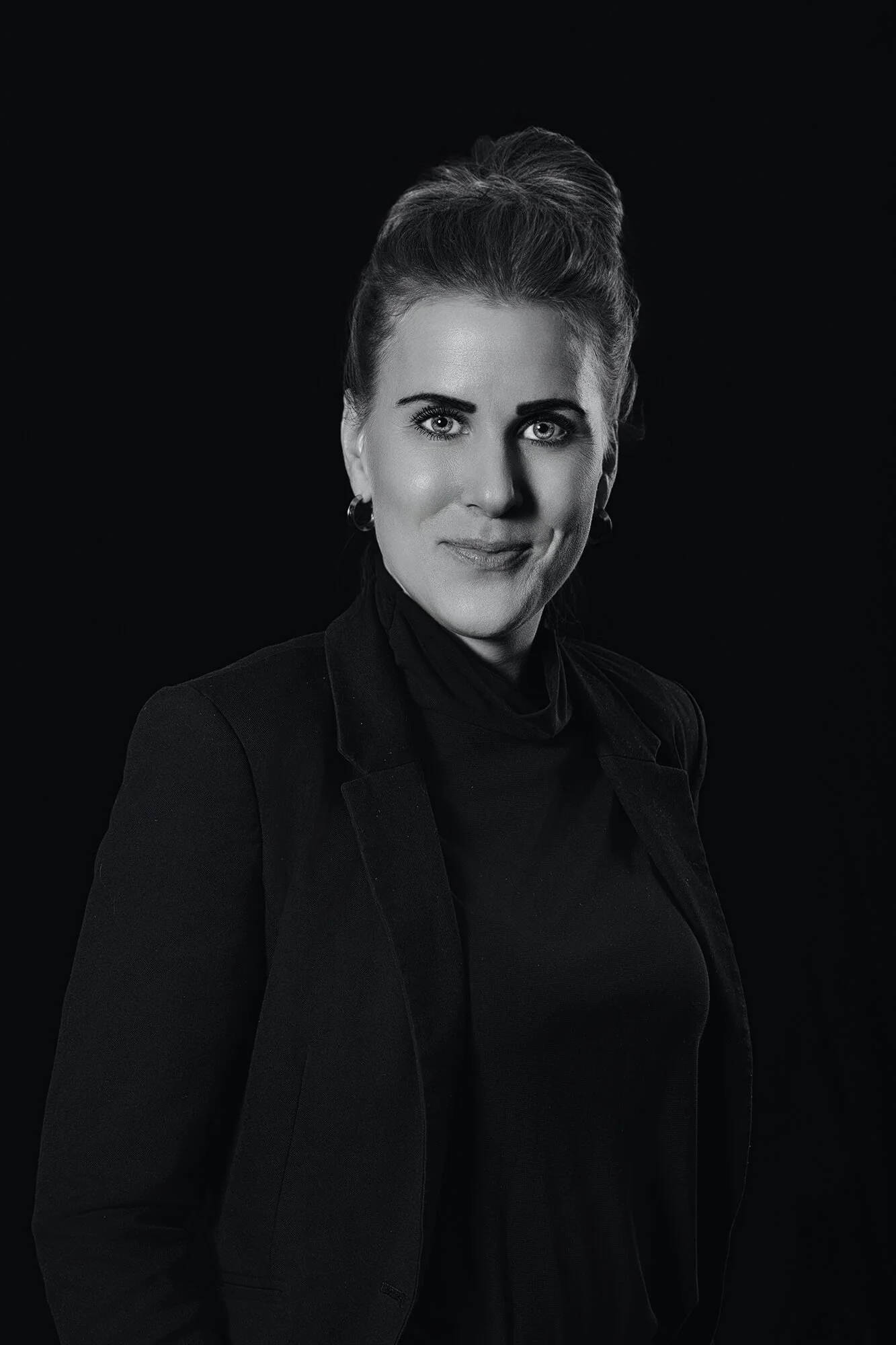 Black and white portrait of a woman with her hair styled up, wearing earrings and a dark blazer over a turtleneck, against a dark background.