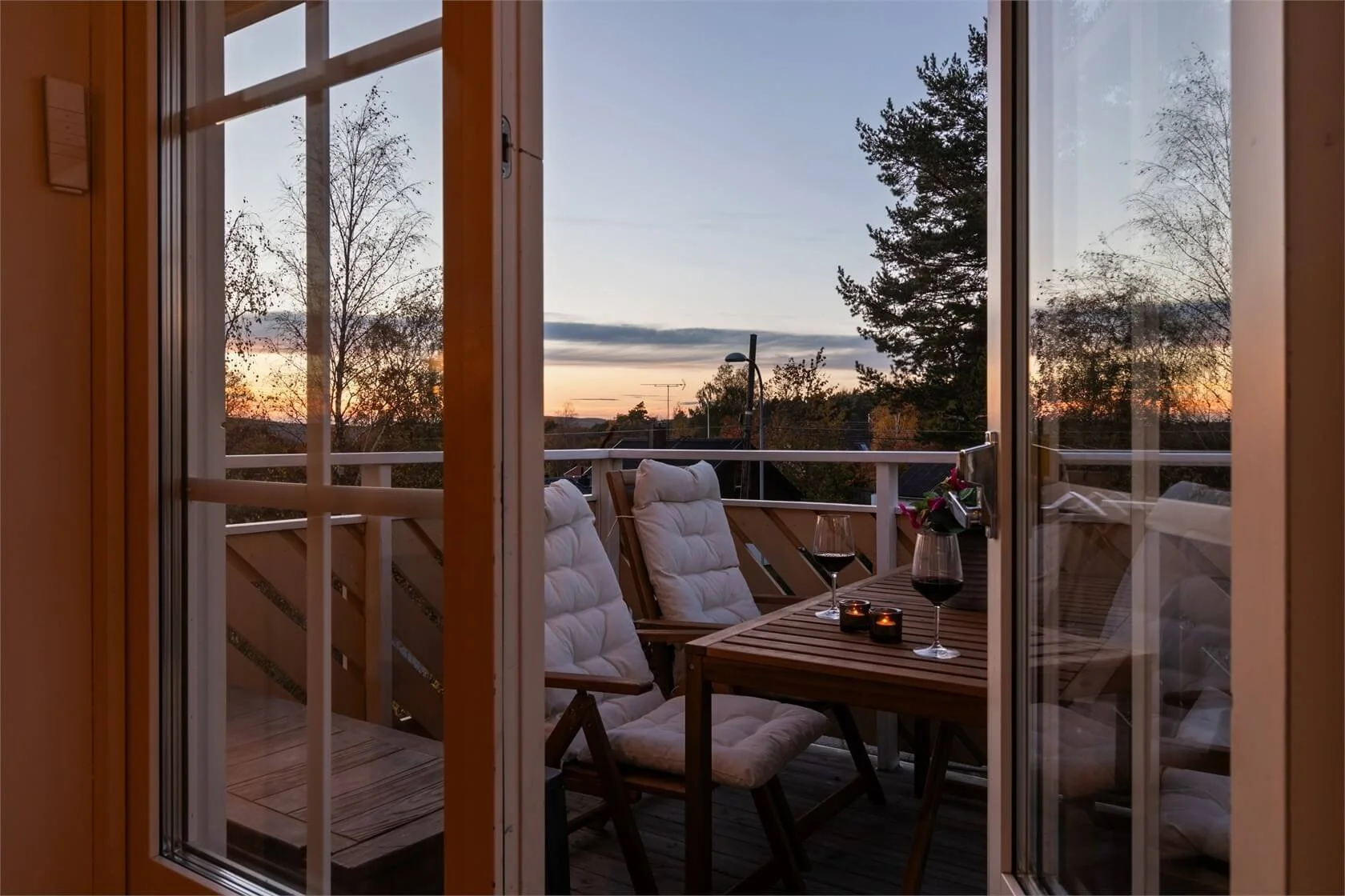 View of a balcony with a wooden table, two cushioned chairs, wine glasses, candles, and a vase with flowers, overlooking trees and a sunset sky.