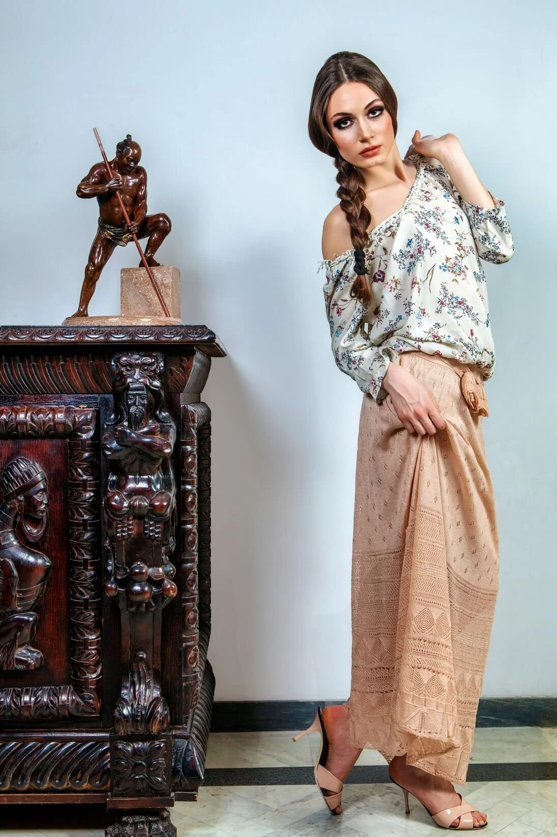 A woman with long brown hair in a braid, wearing a floral off-shoulder top, beige wide-leg pants, and nude high heels, standing next to a dark wood carved cabinet with statues on top, against a light-colored wall.