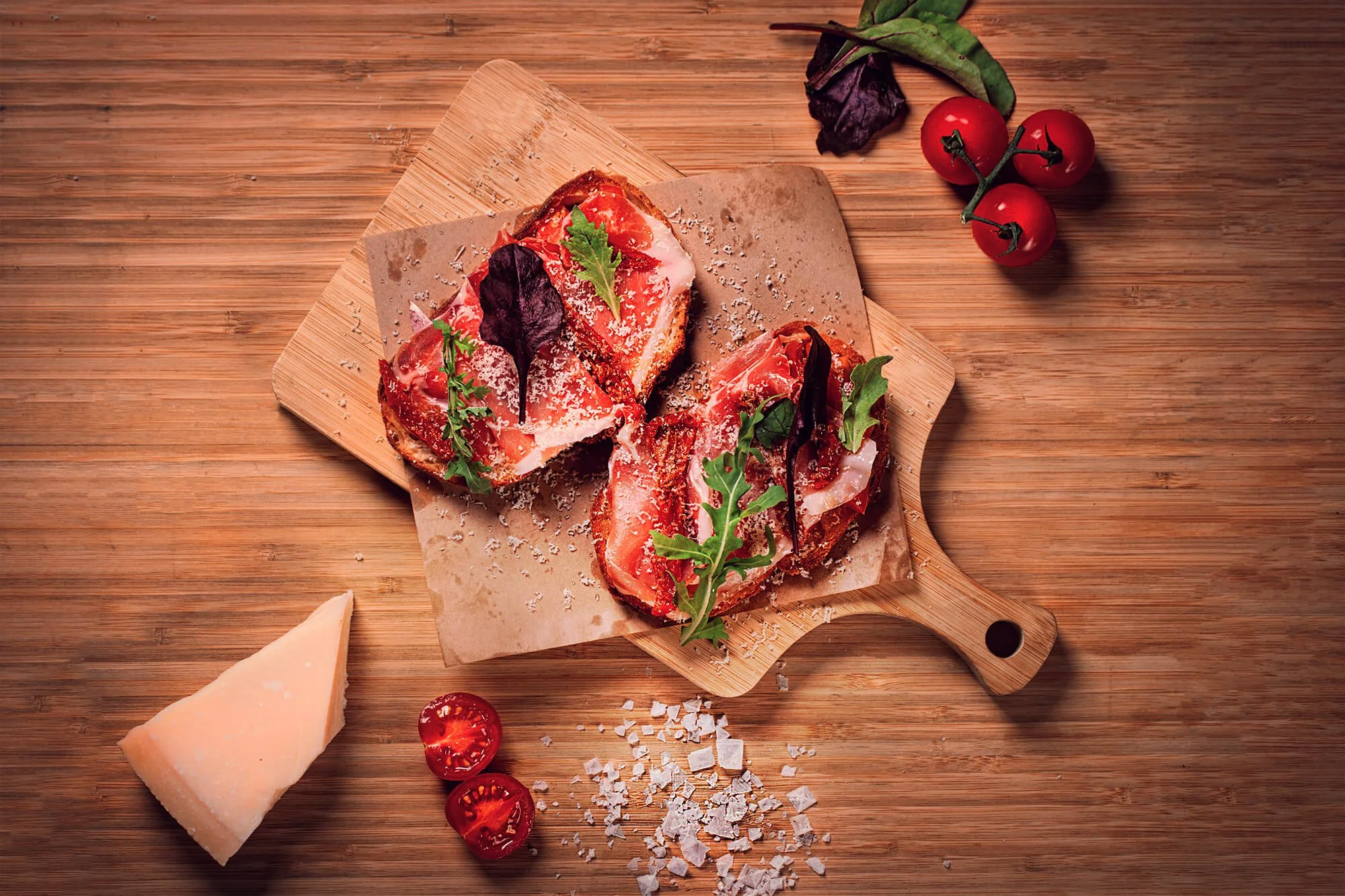 Slices of toasted bread topped with tomatoes, grated cheese, and fresh herbs on a wooden serving board, with cherry tomatoes, cheese, and basil on a wooden surface.