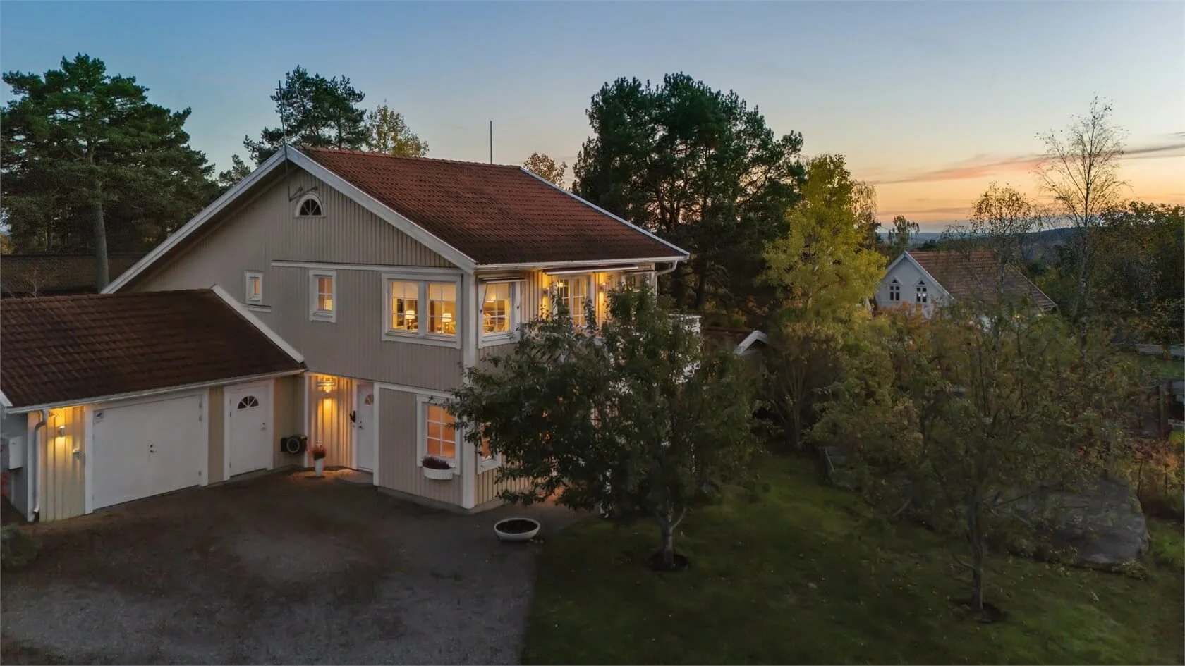 A two-story house with vertical wooden siding, white trim, and a brown roof, with lights on inside, surrounded by green trees at sunset.