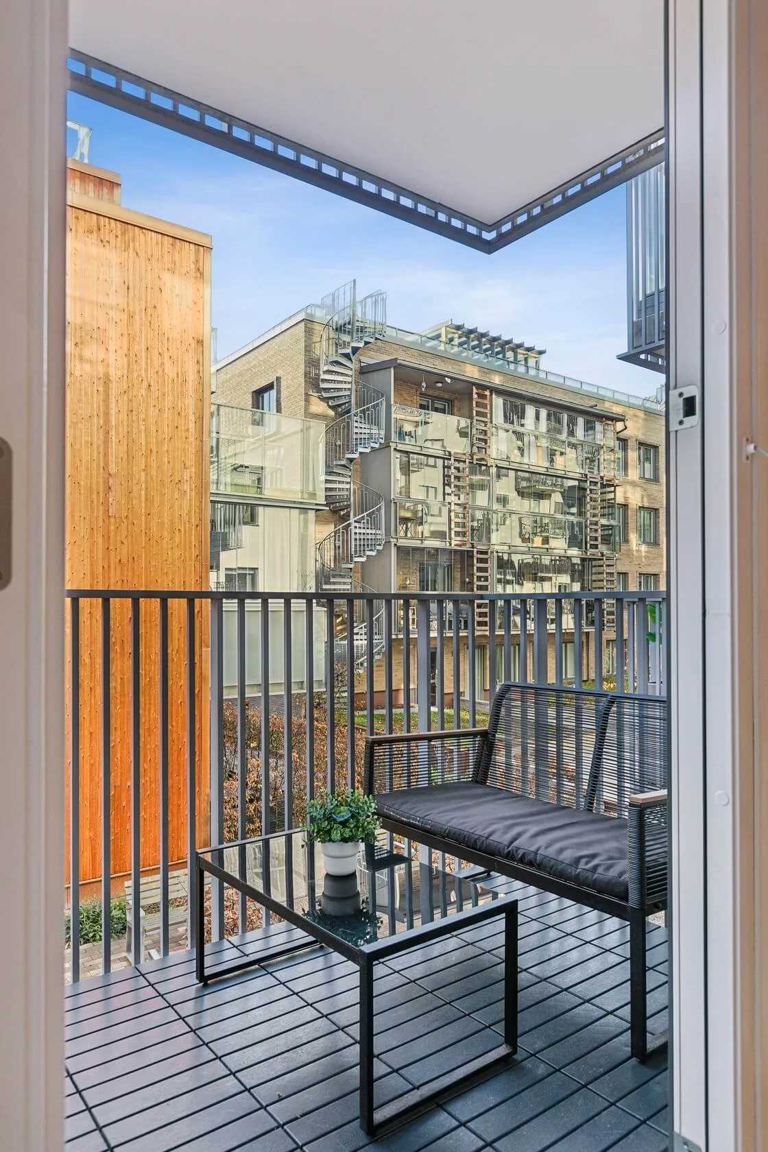 Balcony with black metal and mesh bench, black glass table with a small potted plant, and view of neighboring modern apartment buildings with glass balconies and external spiral staircase.
