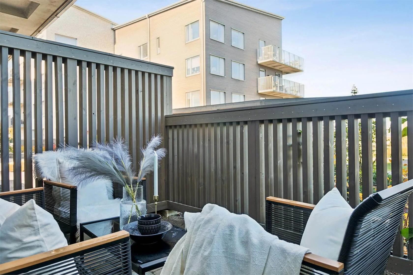 Outdoor balcony with black metal chairs, a small black table, white cushions and throws, decorative pillows, a glass vase with pampas grass, and a tall white candle in a gold holder, with a wooden privacy fence and a multi-story building with balconi