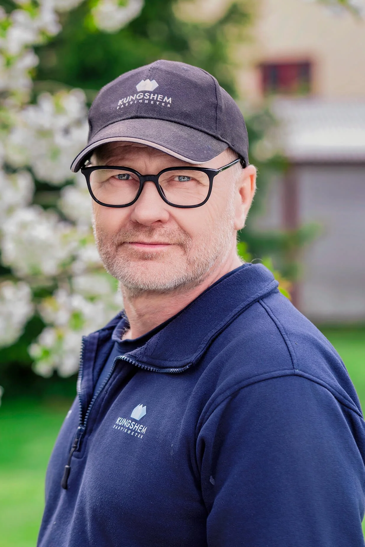 A middle-aged man with glasses and a beard, wearing a black baseball cap and navy blue jacket, standing outdoors with a blurred background of greenery and a building.