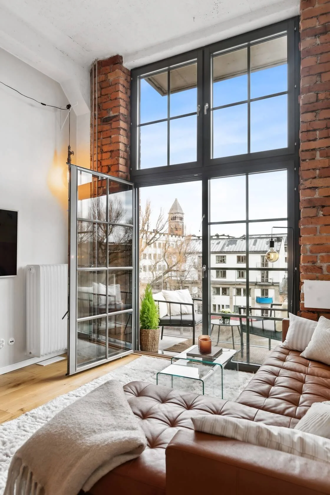 Living room with large industrial-style windows, a brown leather tufted sofa, a white area rug, a glass coffee table, a potted plant, and a balcony with outdoor furniture and a view of a church steeple and city buildings.
