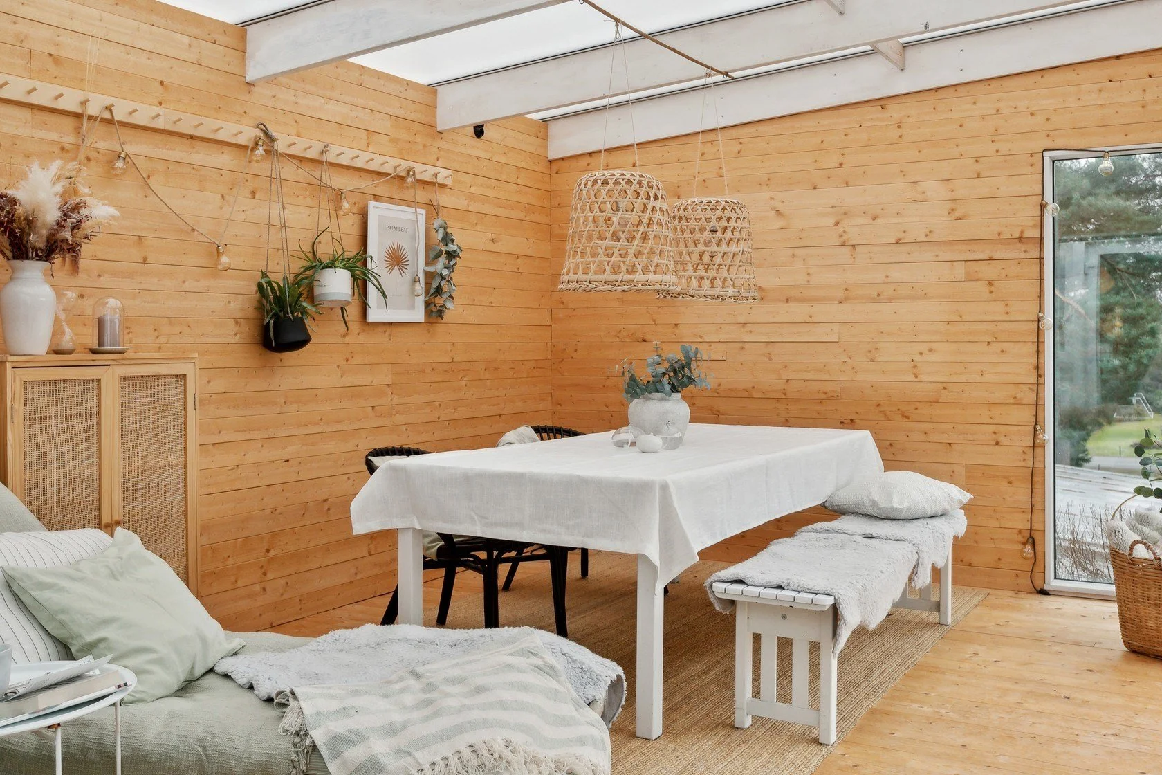 Bright dining area with wooden walls and ceiling, featuring a white table with a tablecloth, flanked by a bench and chairs, decorated with a white vase and greenery, hanging woven light fixtures, and a decorative wall with plants and artwork.