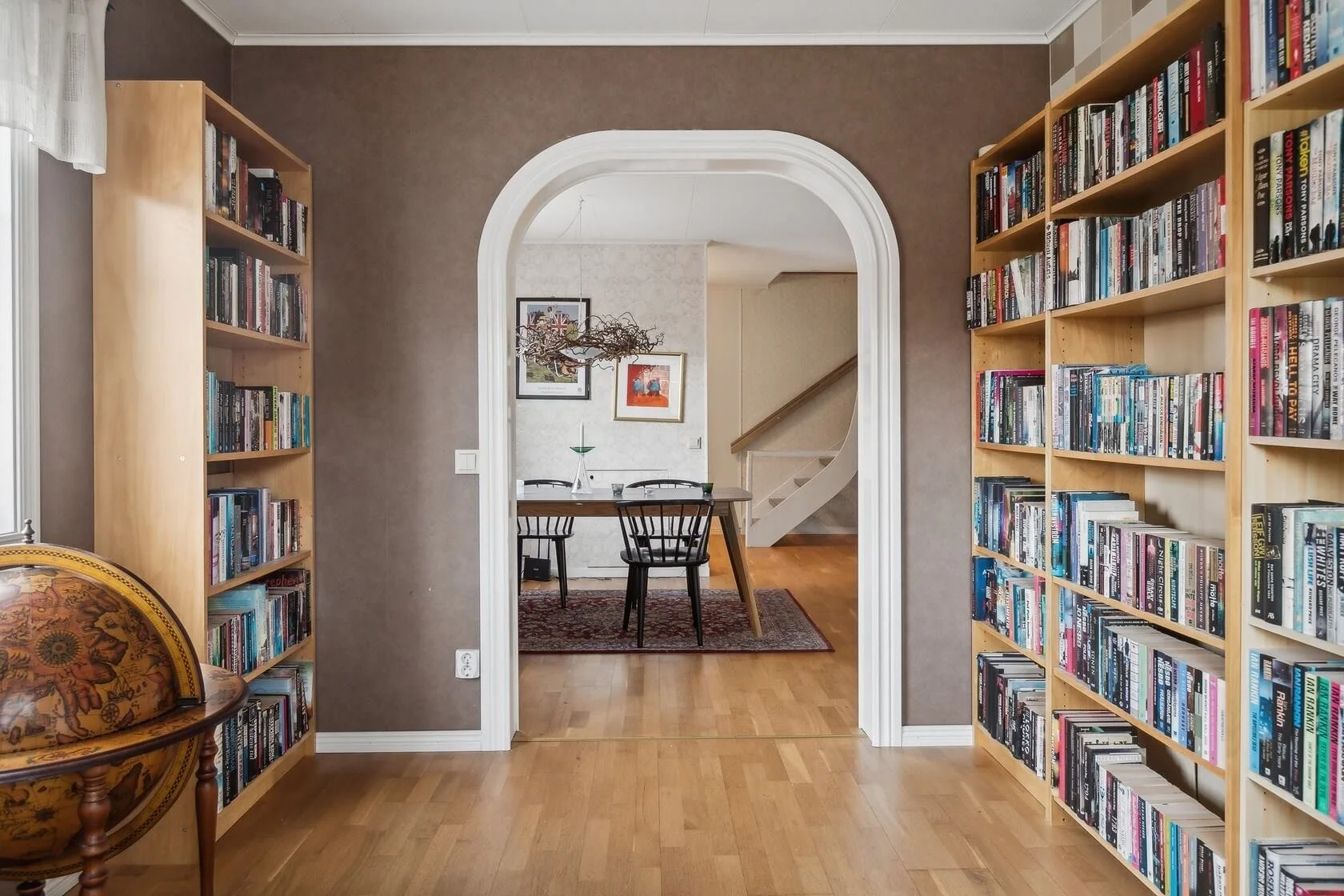 View through an arched doorway into a dining area with a table, four chairs, a rug, framed art on the wall, and stairs in the background. The foreground features two wooden bookshelves filled with books, with a globe on a small table to the left.