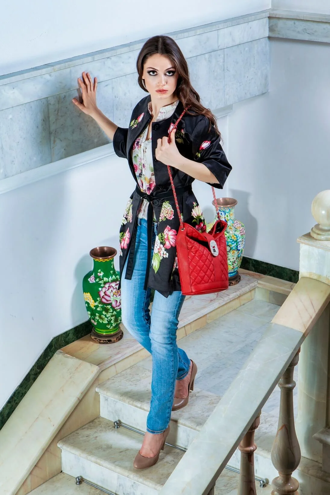 A woman with long brown hair, dressed in a black floral jacket, light blue jeans, and beige high heels, stands on a marble staircase inside a building with decorative vases behind her.