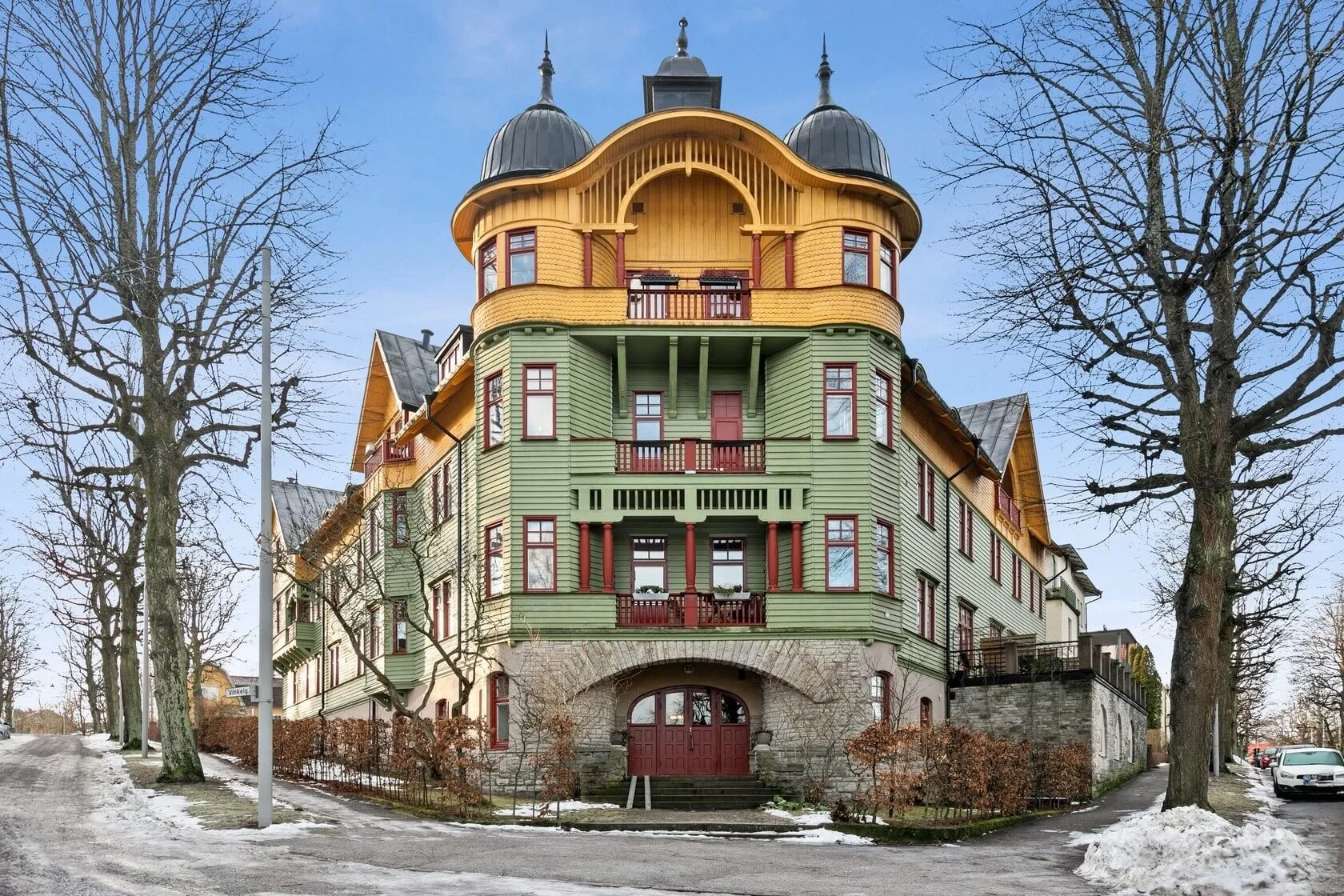 A colorful, multi-story Victorian house with green and yellow wood siding, red accents, and a rounded turret with a domed roof. The house features multiple windows, balconies, and an arched stone entrance on a cold day with snow patches on the ground