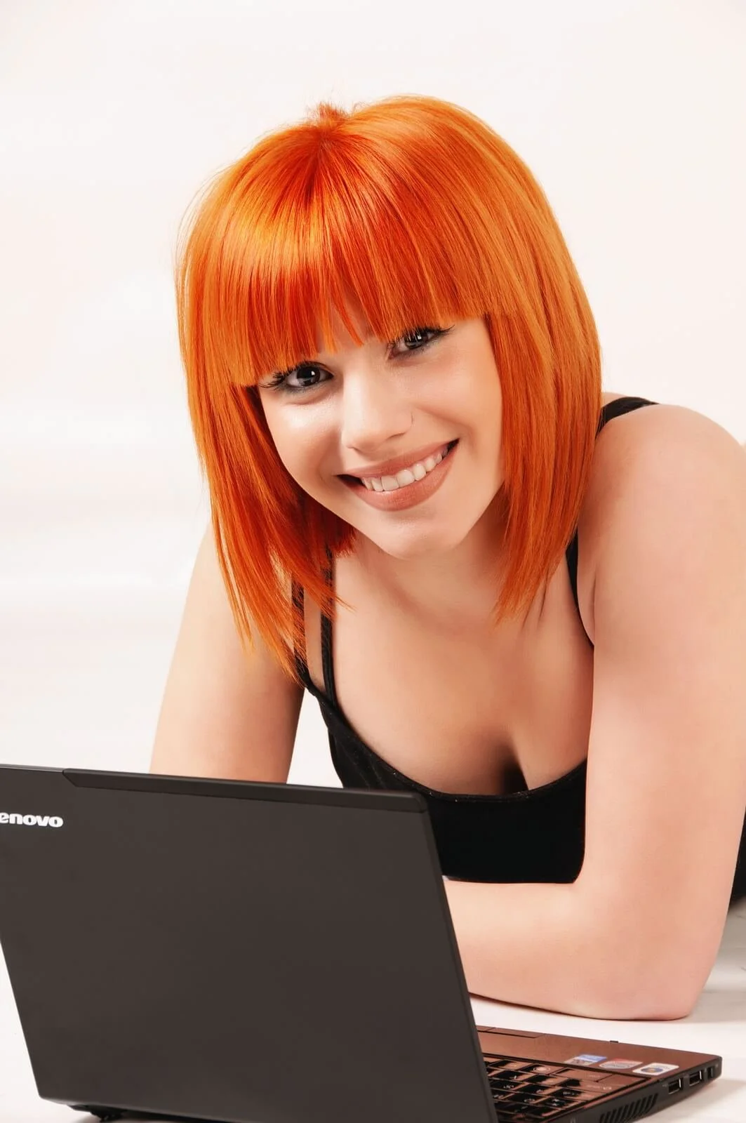 Young woman with bright orange hair smiling at a laptop with a black body, wearing a black top, against a plain white background.