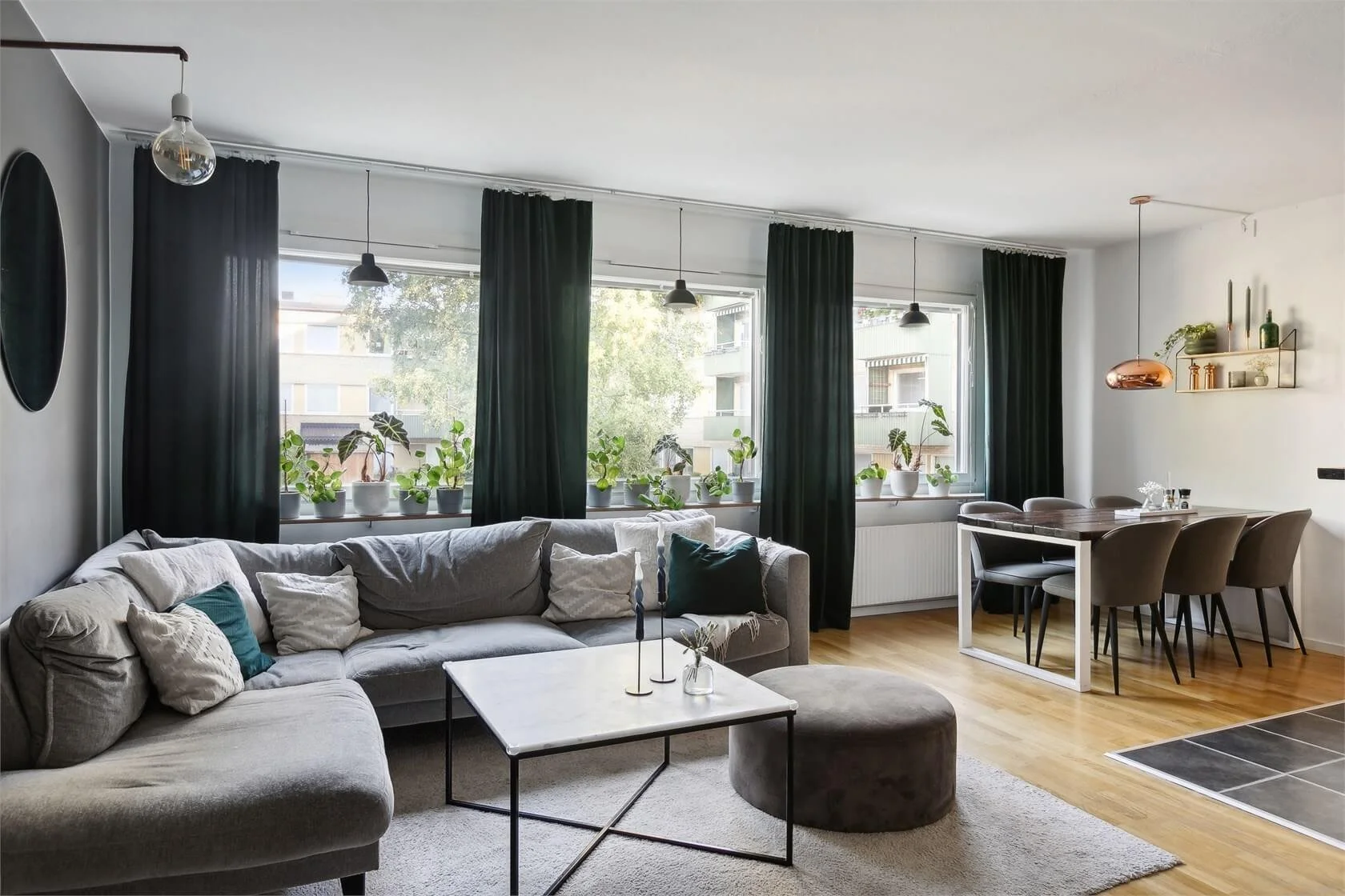 Living room with a gray sectional sofa, pillows, a marble coffee table, and a gray pouf, large windows with black curtains, a dining table with six chairs, and decorative shelves with plants and vases.