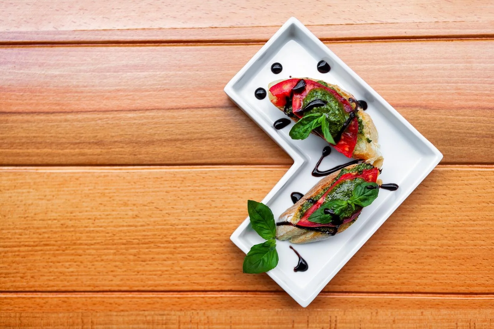Caprese bruschetta with tomato, basil, mozzarella, balsamic glaze, and olive oil on toasted bread on a white rectangular plate placed on wooden surface.