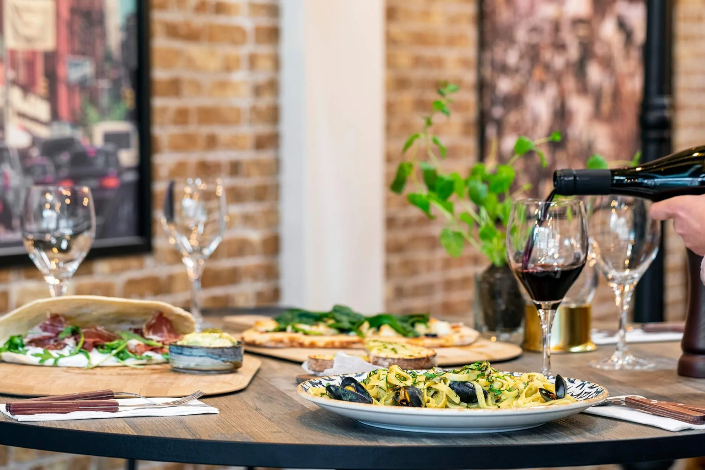 A dining table with glasses of red wine, a plate of mussels and pasta, a pizza, and a charcuterie board, set in a cozy restaurant with brick walls.