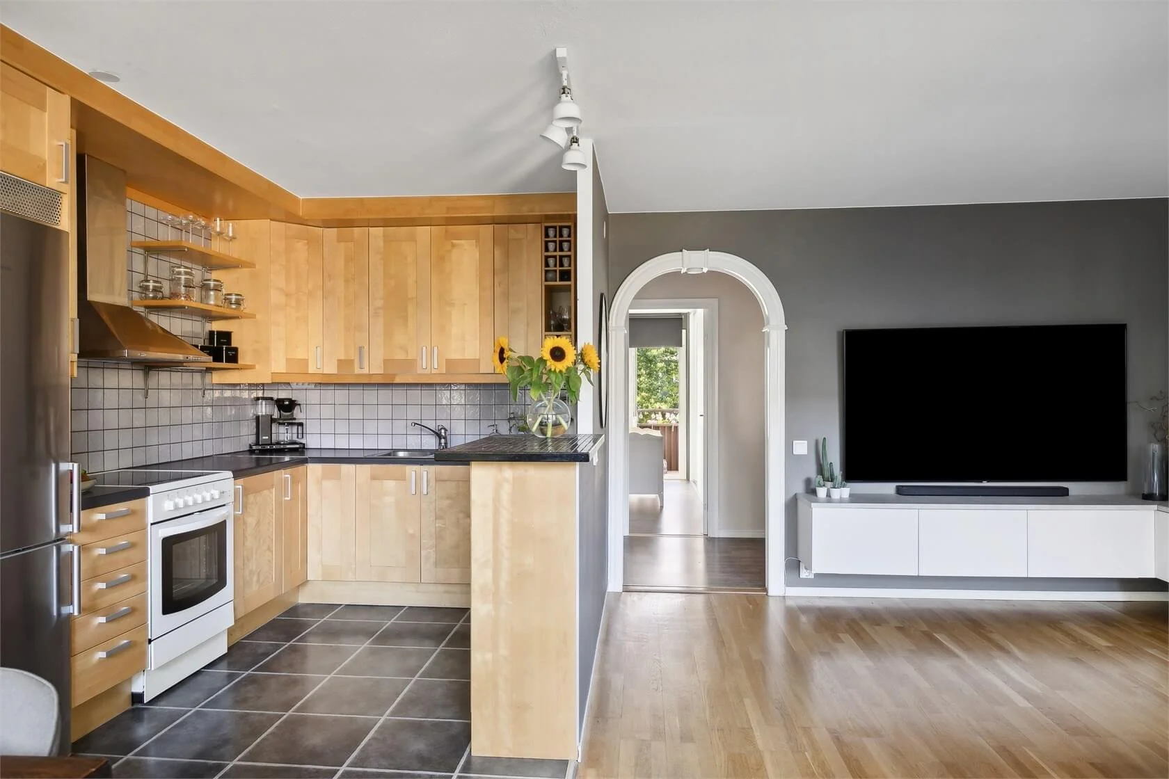 Kitchen with wooden cabinets, black countertop, white tile backsplash, stainless steel refrigerator, white stove, coffee maker, and sunflower flower arrangement on the counter. Connected to a living room with a large flat-screen TV and potted plants,