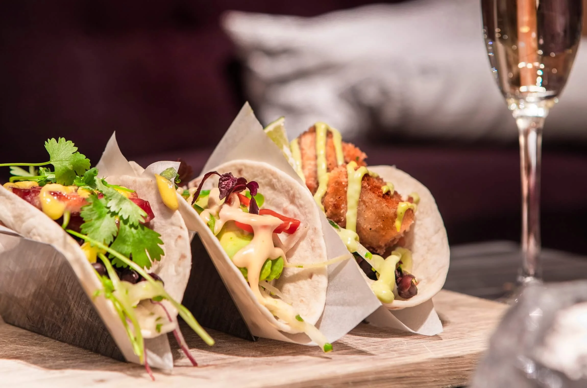 Three tacos topped with fresh cilantro, colorful vegetables, and drizzle of yellow and white sauces on a wooden board, with a glass of sparkling wine in the background.