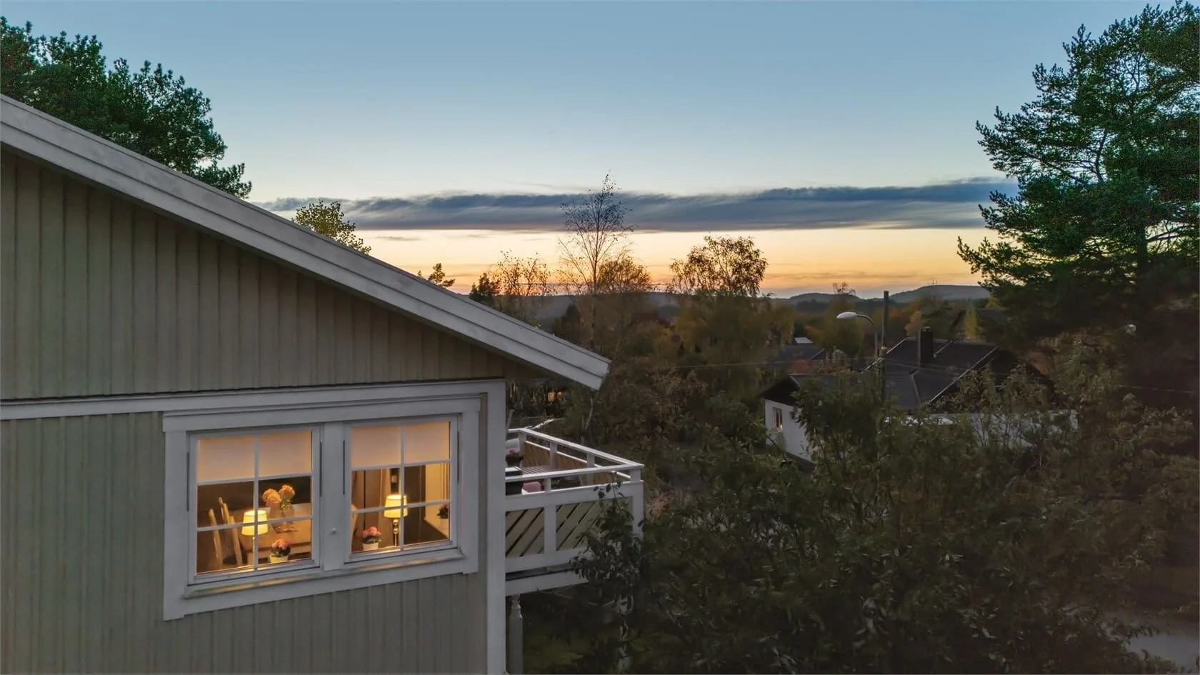 View of a house exterior at sunset with the interior lit, showing a window with lamps and flowers, surrounded by trees and a scenic horizon.