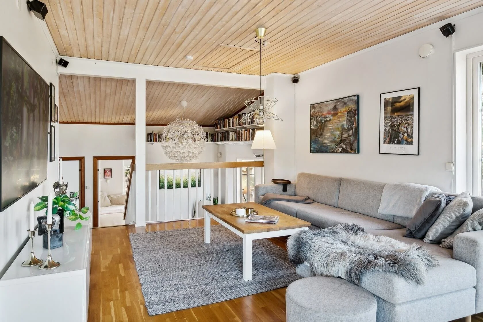 A cozy living room with a gray sectional sofa, a wooden coffee table, and wall art. There are hanging light fixtures, a carpet, and a white media console with candles and a plant. The room features a wooden ceiling and white walls, with a view of an 