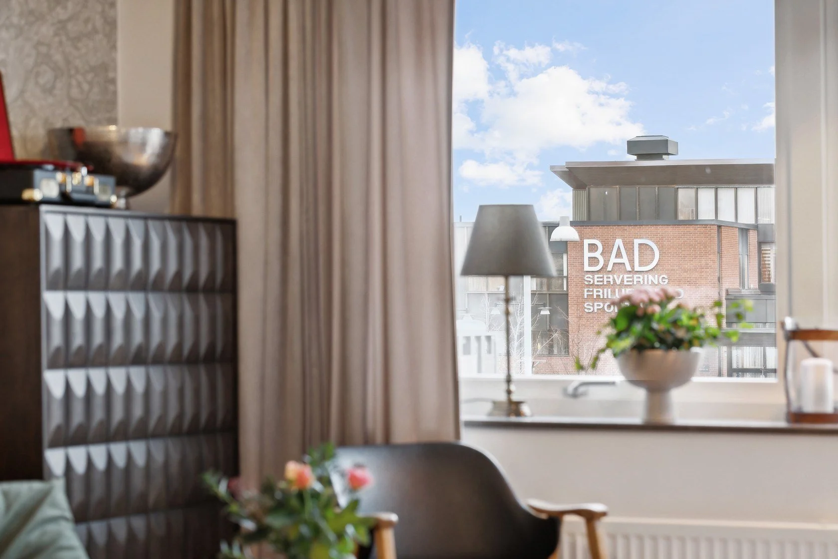 Living room with a window, curtain, lamp, plant, and a view of a building with a sign that reads "BAD SERVING FRUITS, SPORT".