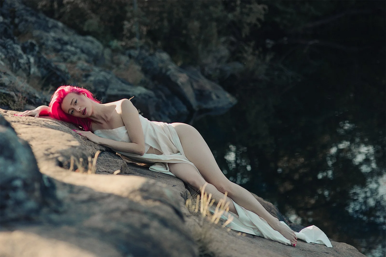 A woman with pink hair lying on a rock by a body of water, wearing a white dress.