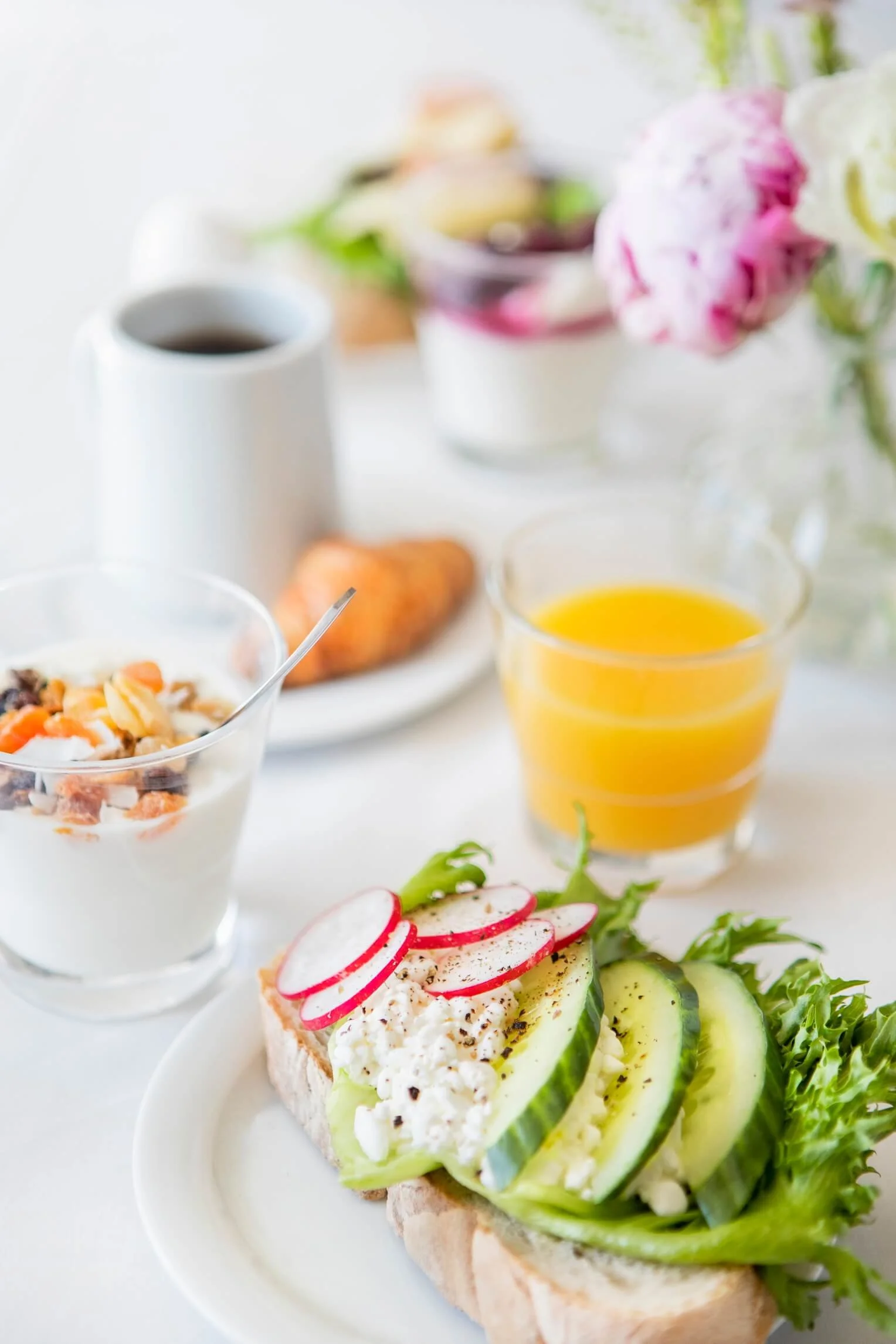 A breakfast spread featuring an open-faced sandwich with cucumbers, radishes, and cheese on bread, a glass of orange juice, a bowl of yogurt topped with nuts, a cup of coffee, and pink and white flowers in a vase.