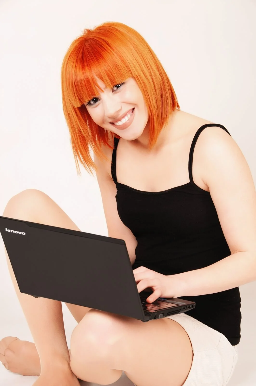 A young woman with bright orange hair, wearing a black tank top and white shorts, sitting cross-legged with a laptop on her lap, smiling at the camera.