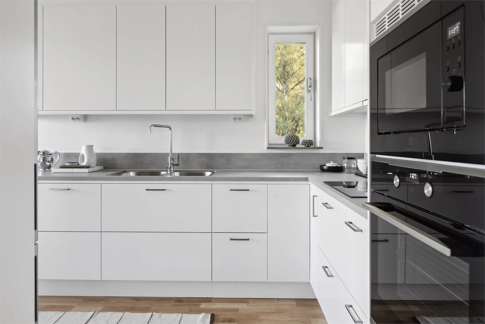 Modern white kitchen with gray countertops, a double sink, and a window showing trees outside. There are black appliances, decorative objects on the window sill, and a white and gray striped rug on wood flooring.