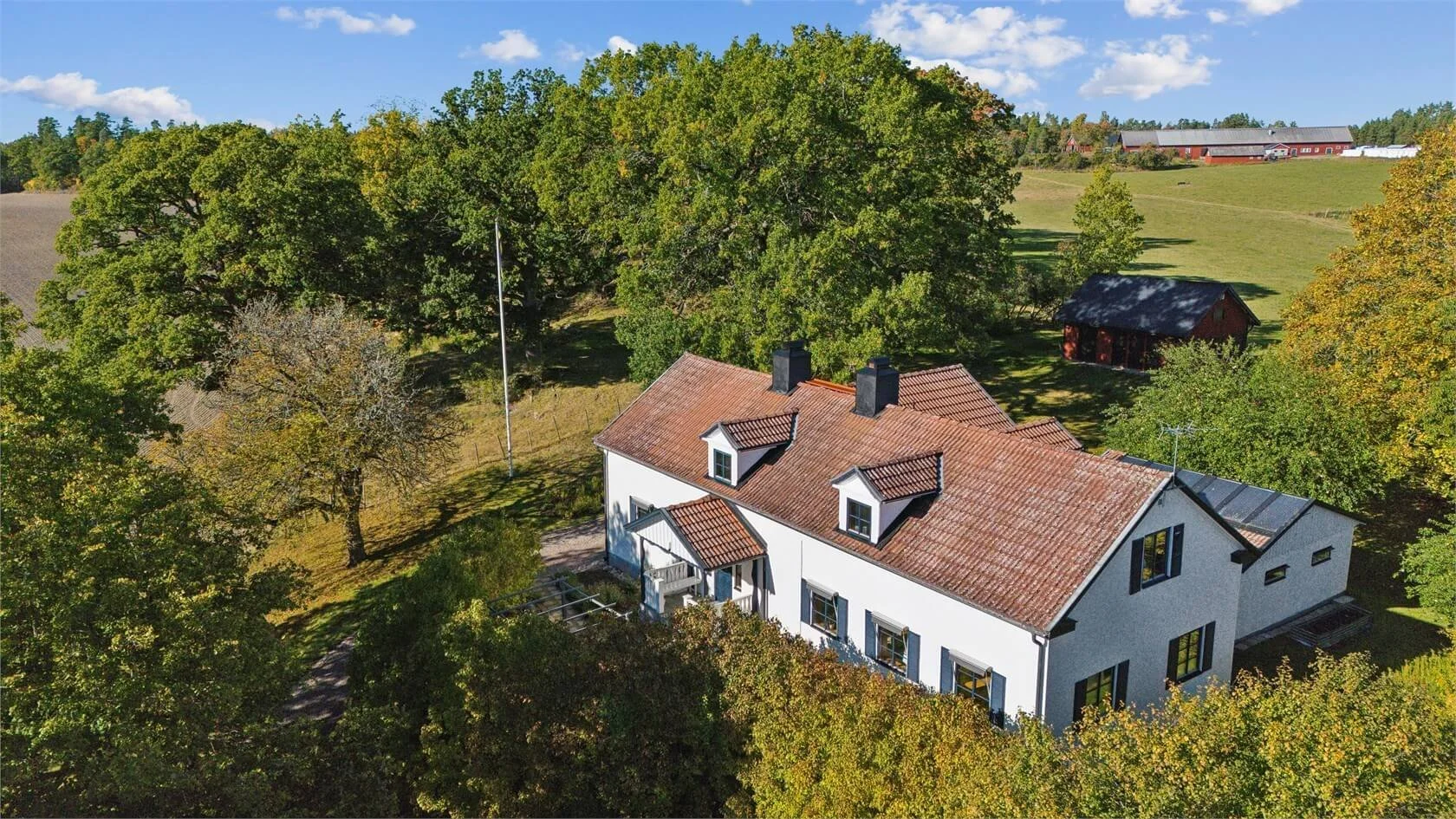 Aerial view of a white house with a red tiled roof, surrounded by green trees and open land, with a clear blue sky overhead.