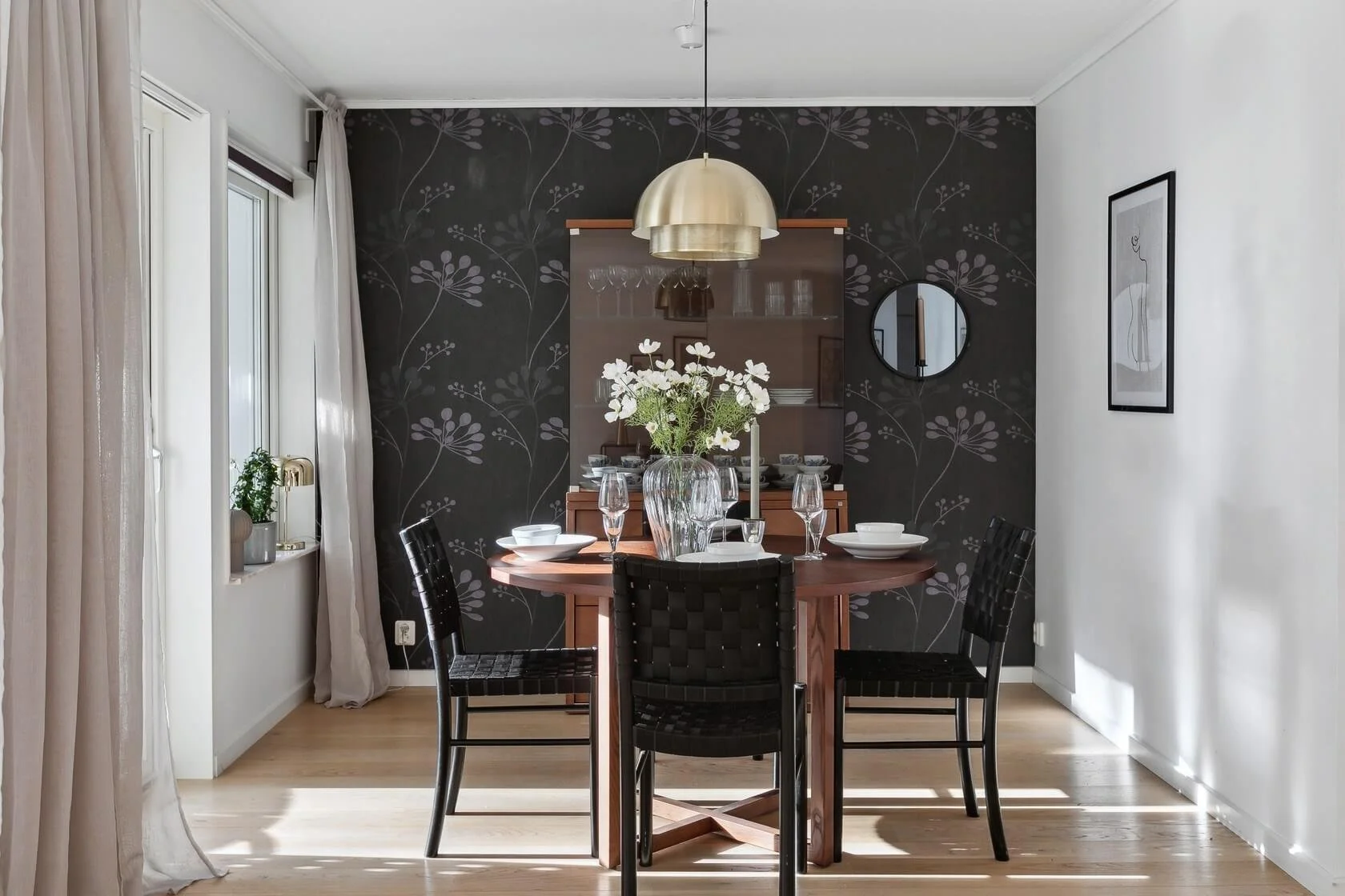 A dining room with a round wooden table set with white dishes, glasses, and a large flower arrangement, four black woven chairs, a black floral accent wall, a gold pendant light, and a framed picture on the white wall.