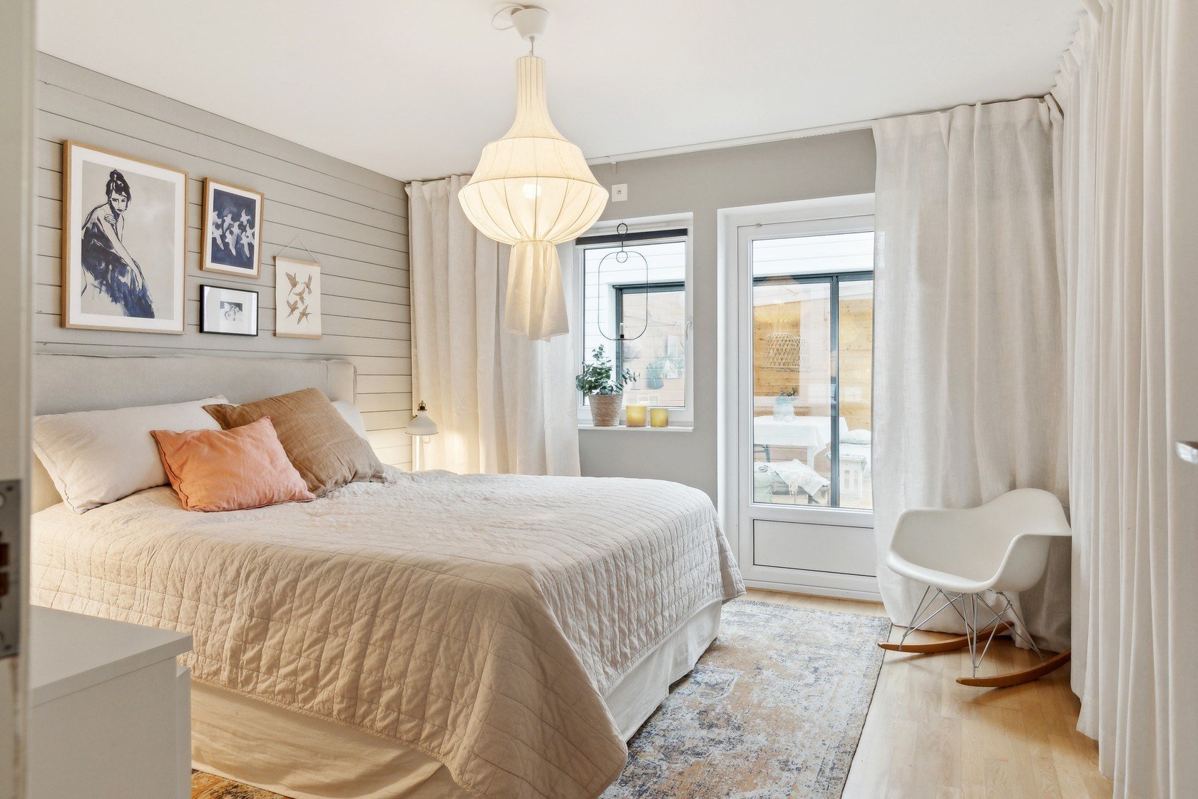 Bright bedroom with white bedding, pink and beige pillows, wall art, a hanging light, window with white curtains, sliding door to a balcony, and a white rocking chair.