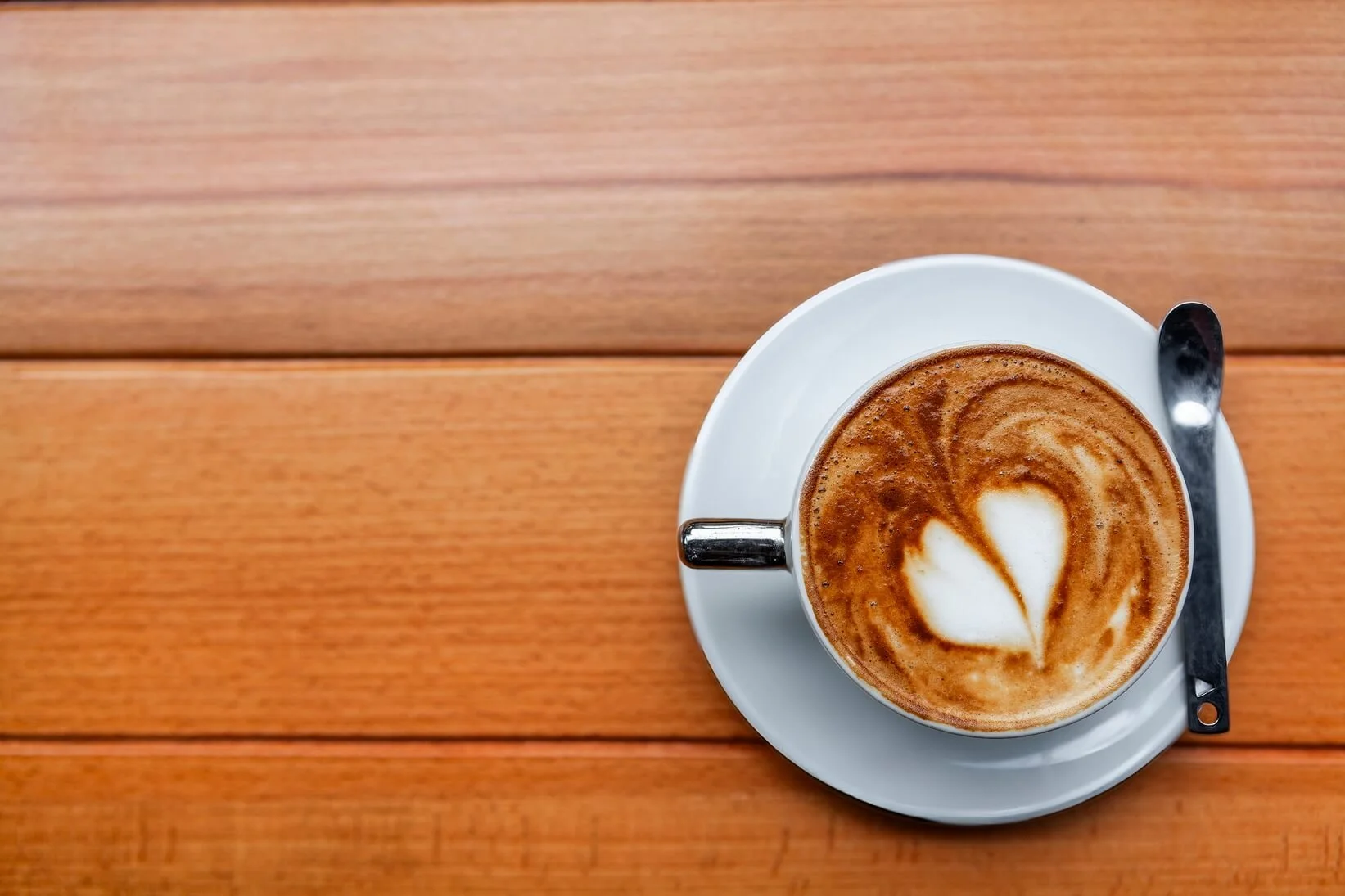 A cup of cappuccino with latte art on top, placed on a white saucer with a spoon, on a wooden table.