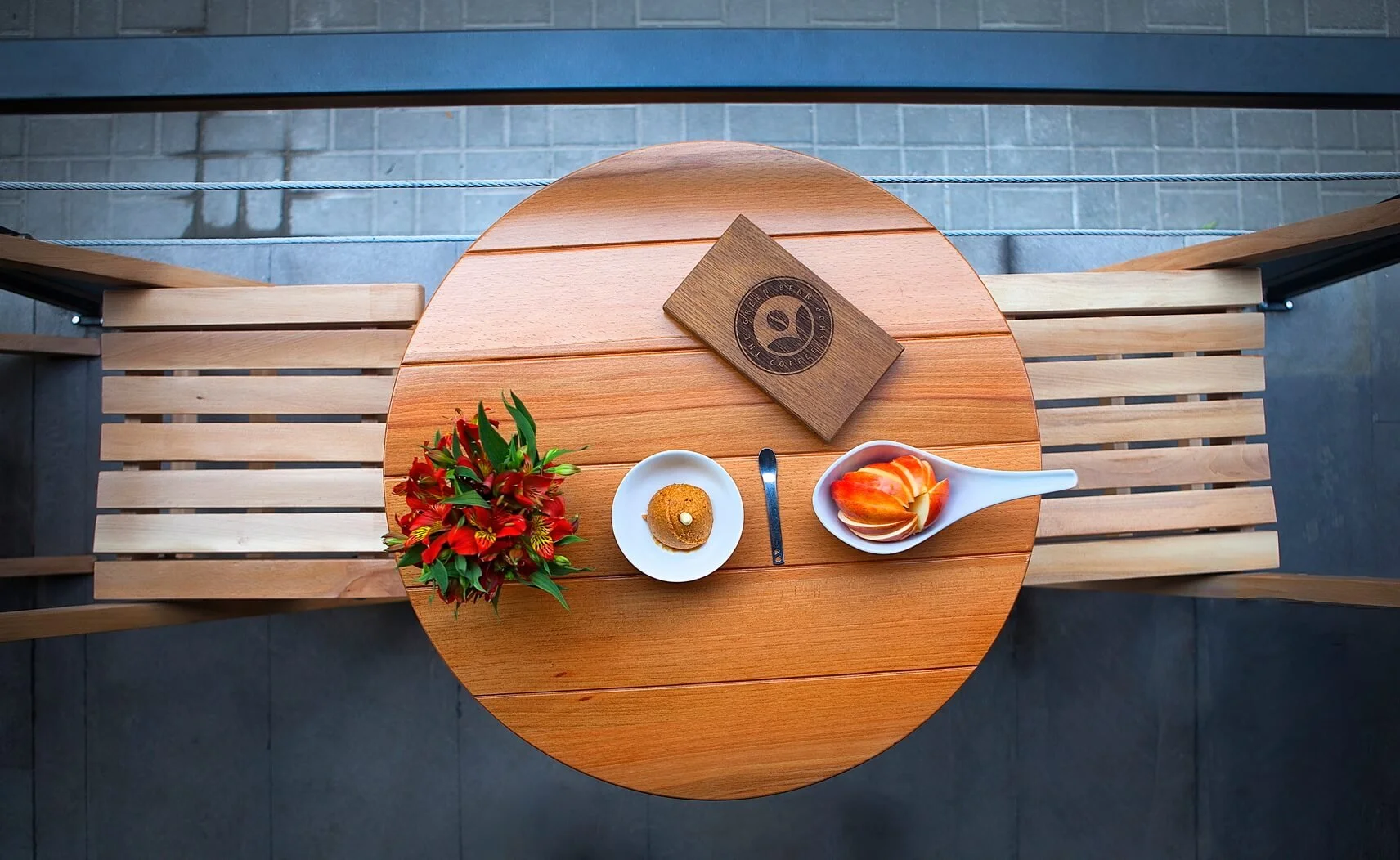 Top-down view of a round wooden table with a flower arrangement, a small plate with a pastry, a spoon, a white container with slices of fruit, and a wooden box with a circular logo.