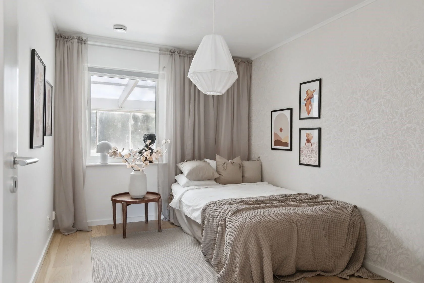 A cozy bedroom with neutral tones, a neatly made bed, framed artwork on the wall, a round wooden side table with a white vase and dried flowers, and a window with sheer white curtains and beige drapes.
