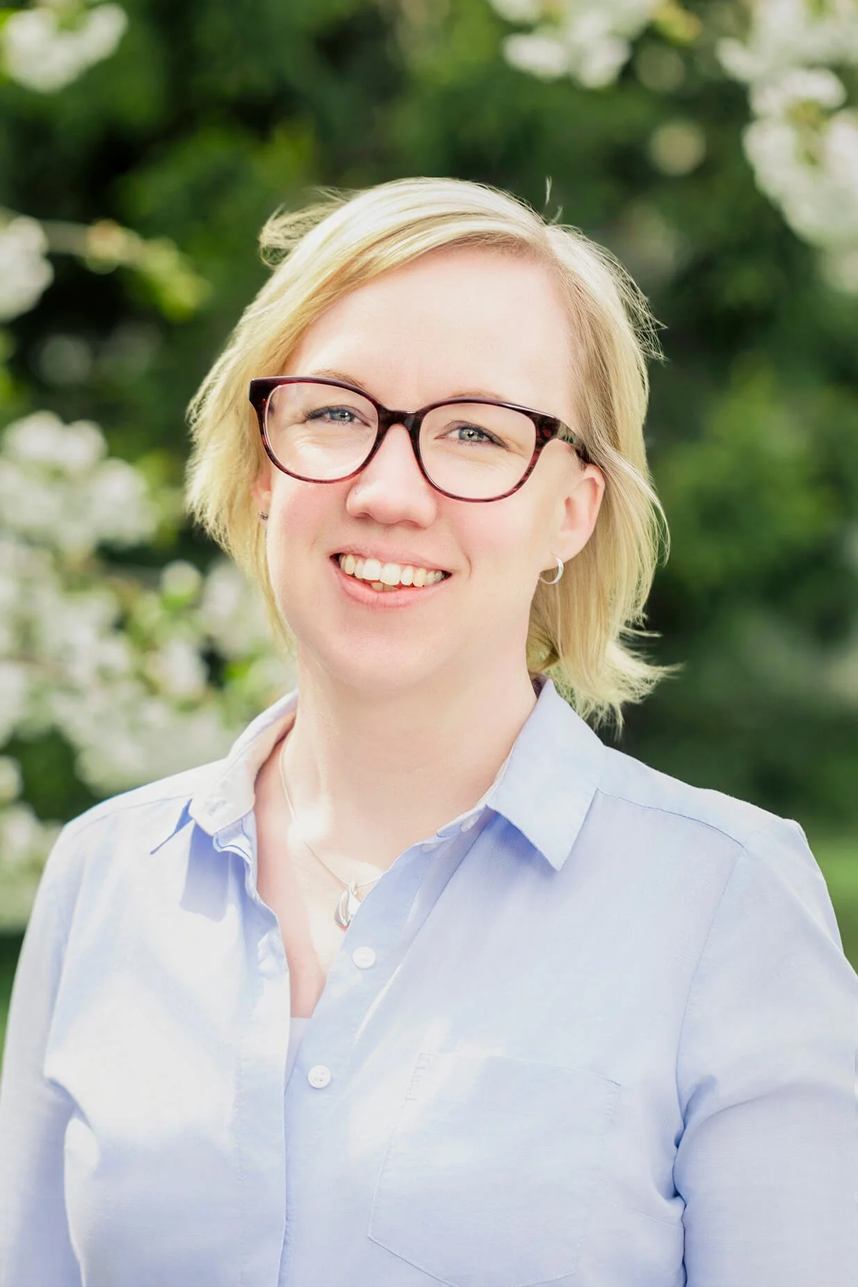 A woman with short blonde hair and glasses smiling outdoors, wearing a light blue button-up shirt.