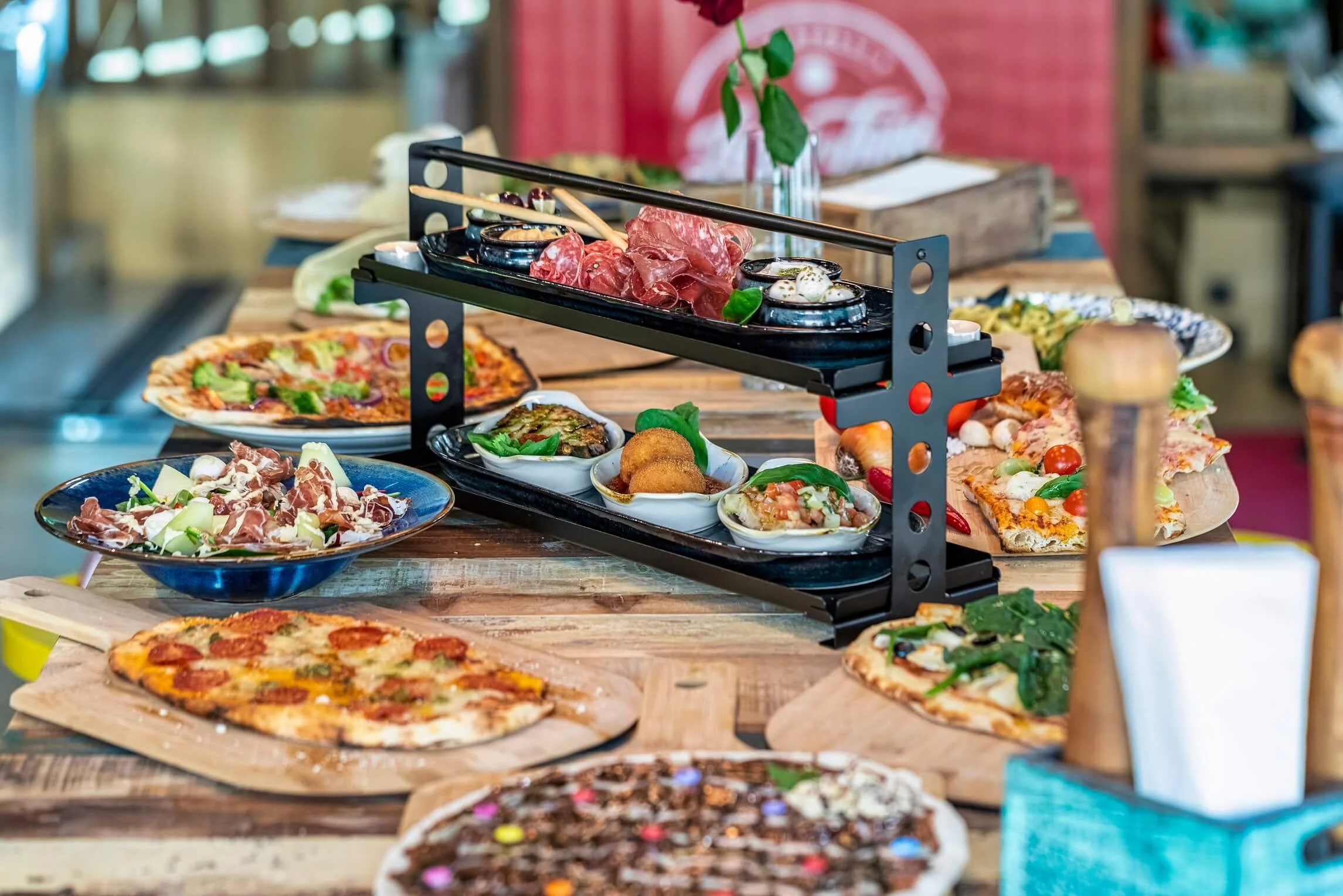 Various dishes on a wooden table, including pizza, pasta, and appetizers, with a tiered serving tray holding meats and condiments.